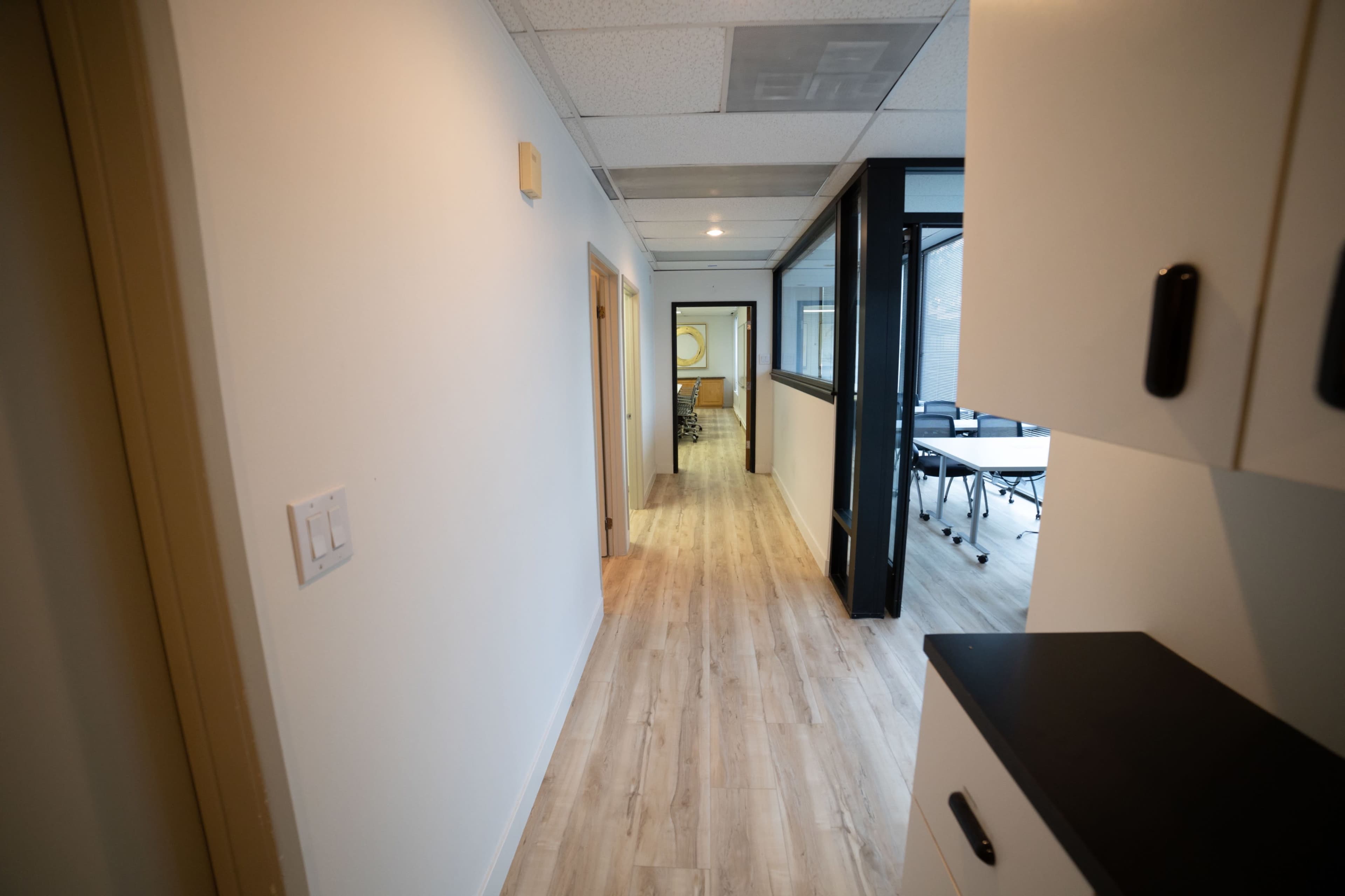 A hallway leads to a glass-walled office space with wooden flooring and neutral-colored walls.