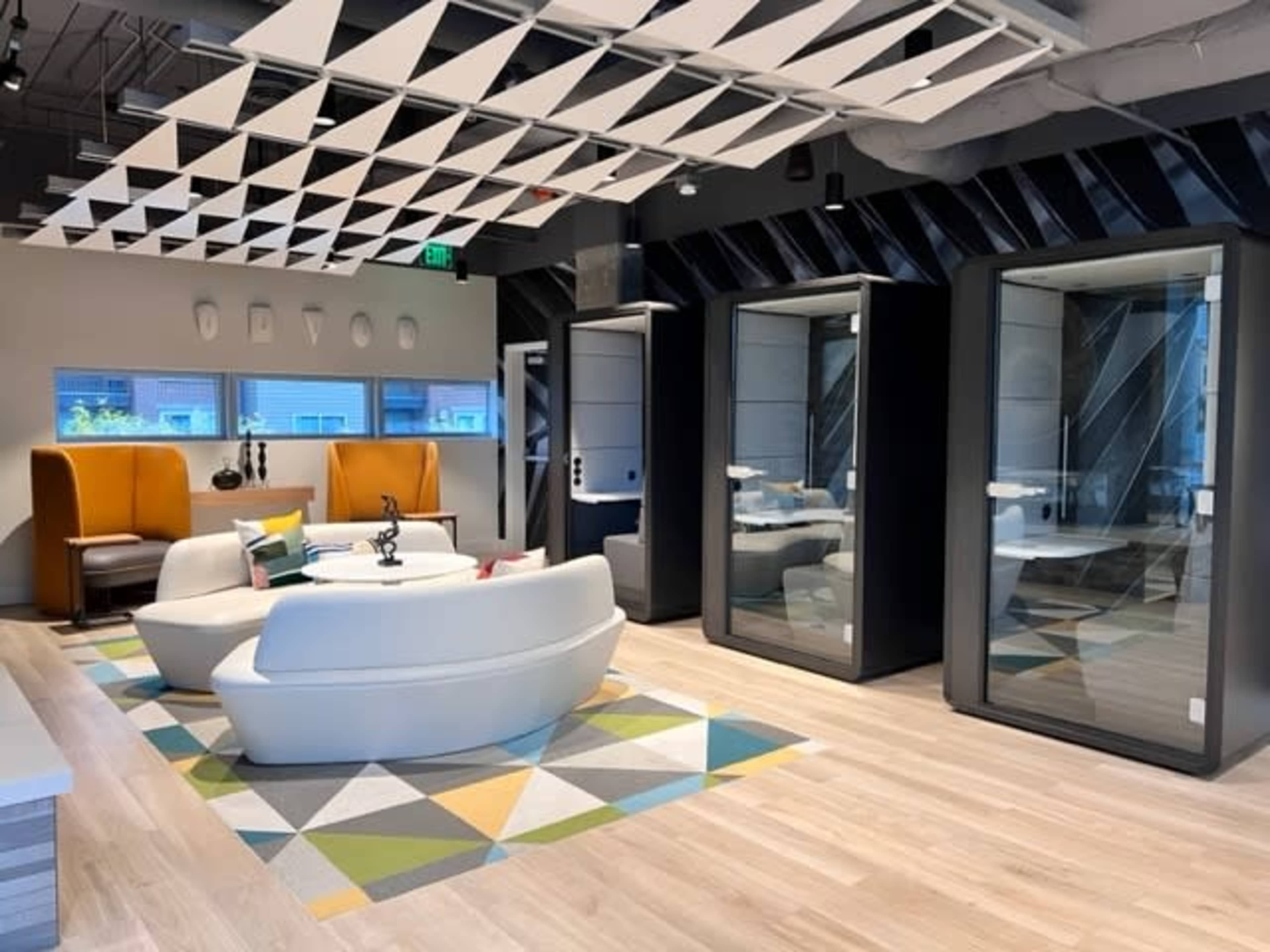 A modern office space featuring a seating area with a white sofa and colorful rug, along with glass-enclosed meeting pods on one side.