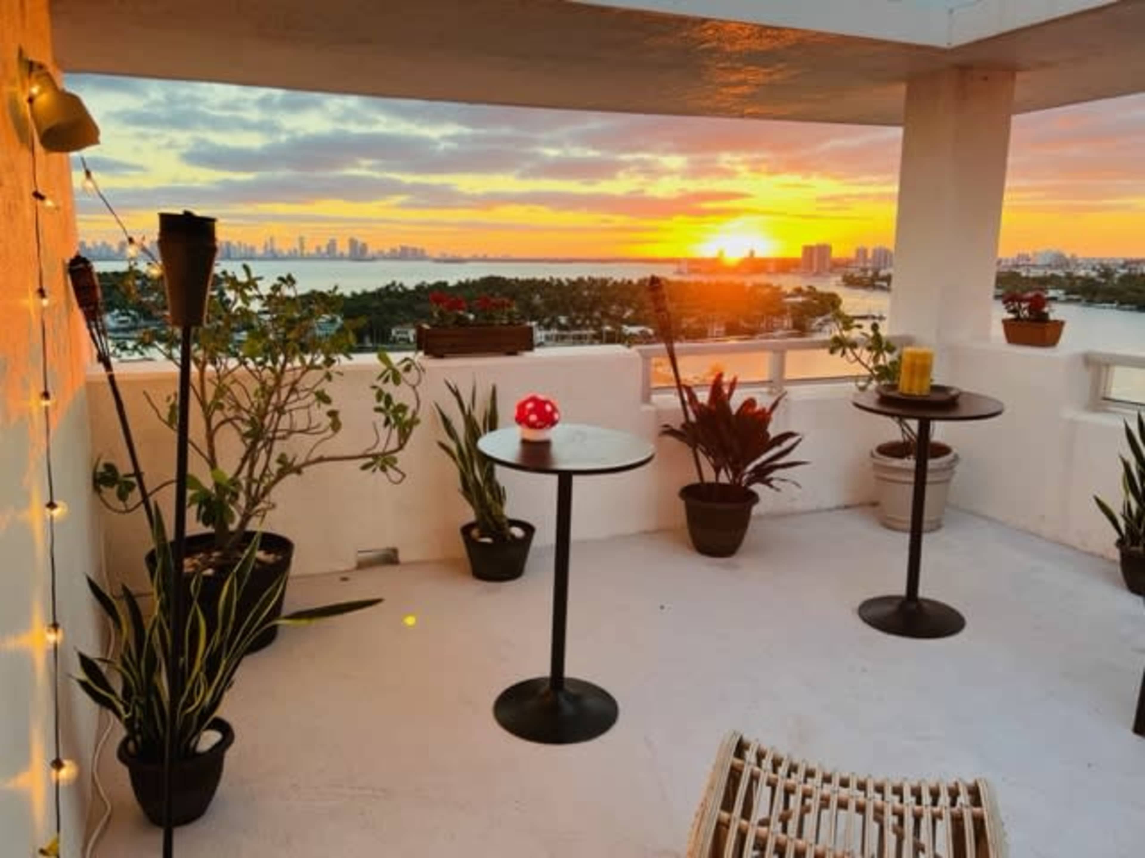 A balcony features two small tables surrounded by potted plants, with a sunset over the skyline in the background.