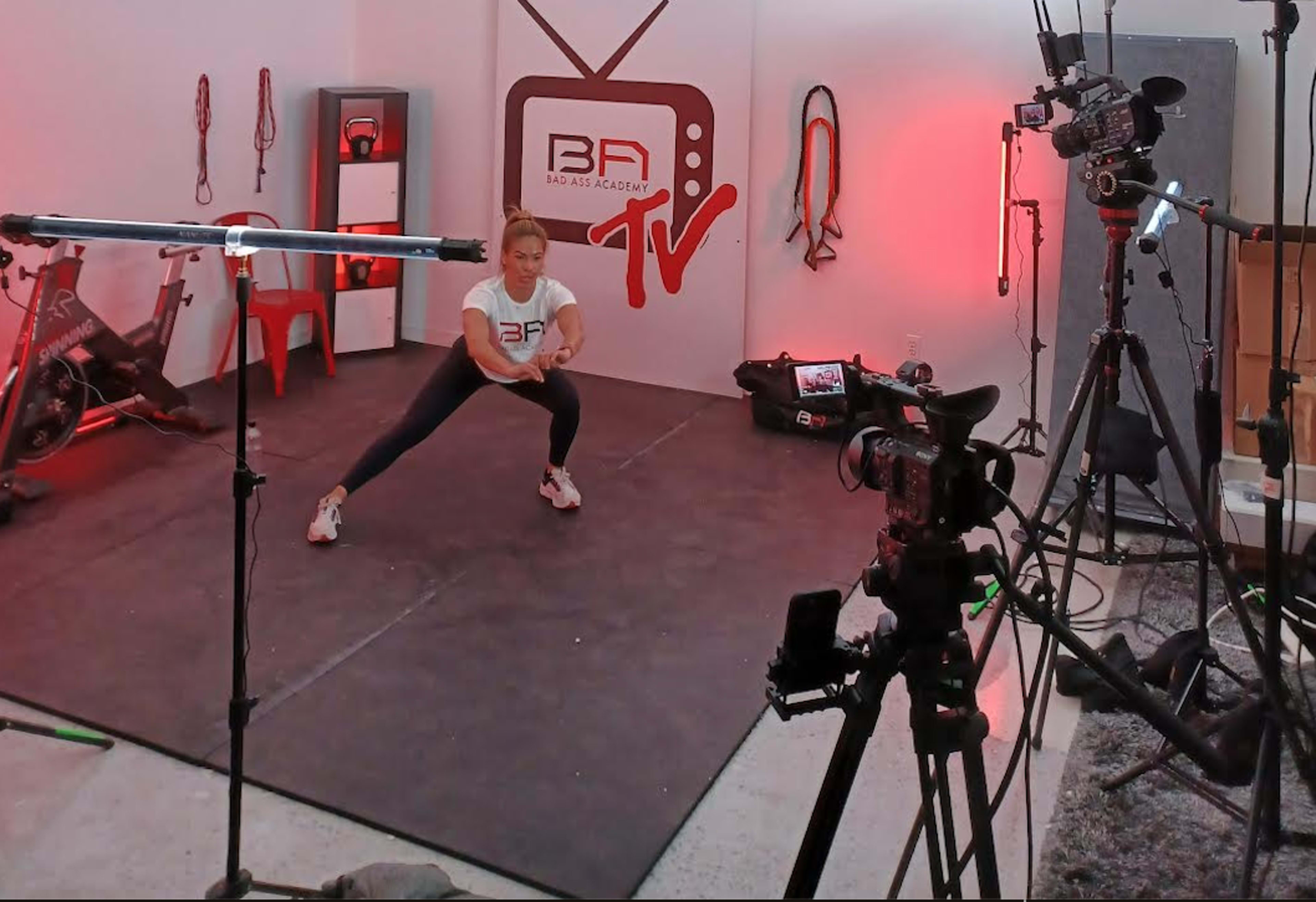 A fitness instructor performs an exercise in a studio equipped with cameras and workout equipment.