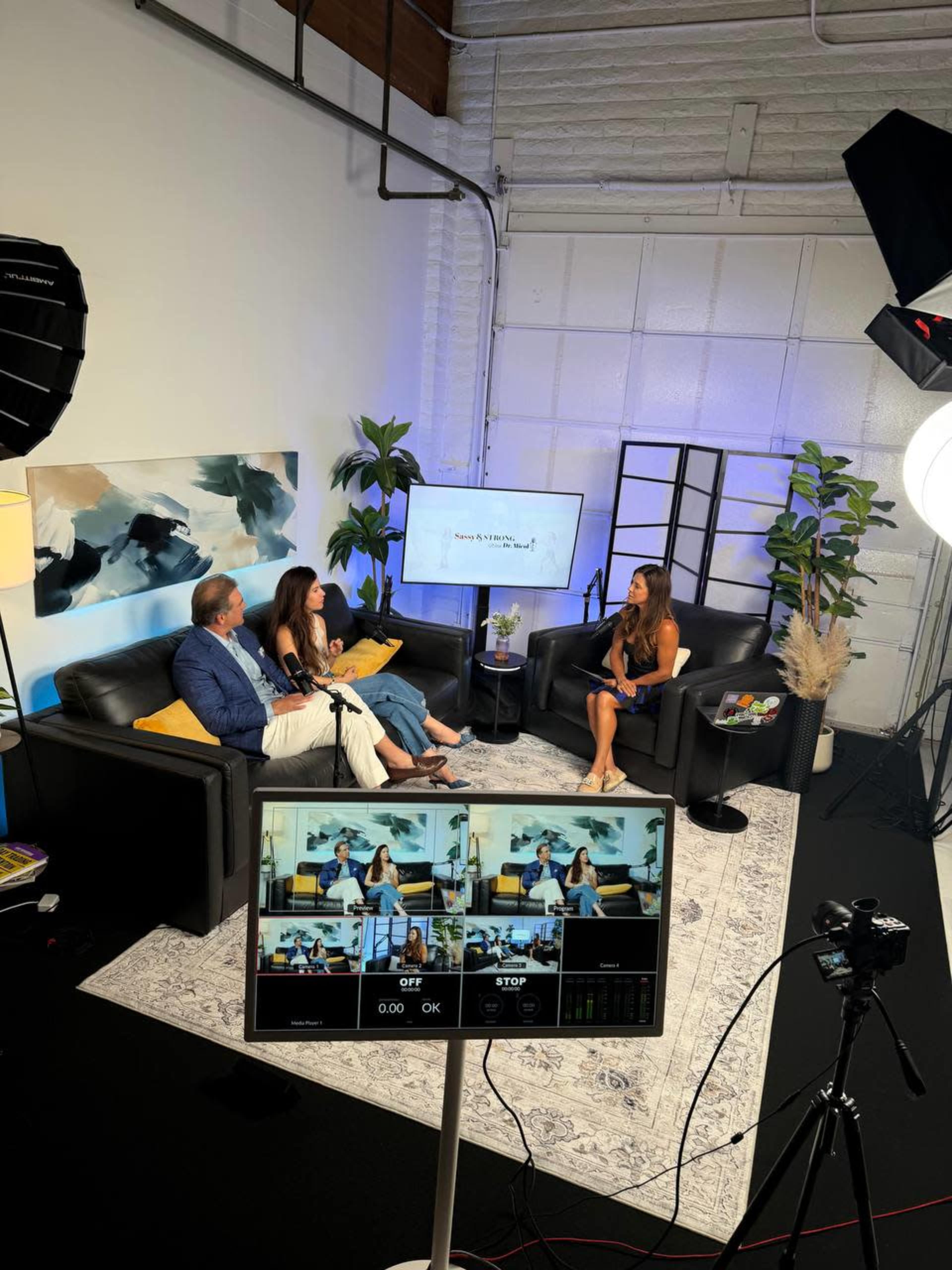 A studio setting features three people engaged in a discussion while seated on black couches, with cameras and lighting equipment positioned for filming.