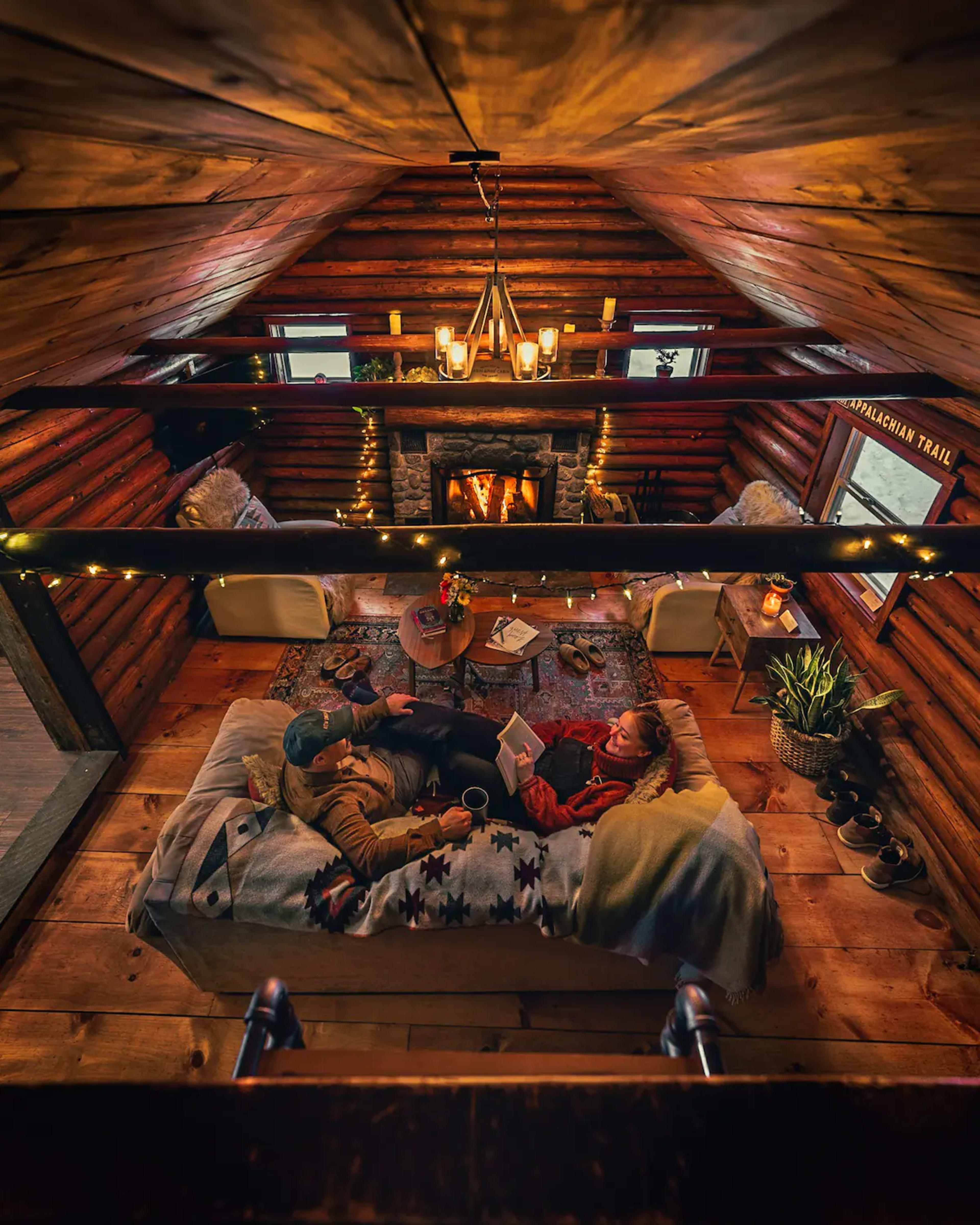A couple relaxes on a cozy couch in a cabin, surrounded by wood-paneled walls and a glowing fireplace, with warm lighting and plants nearby.