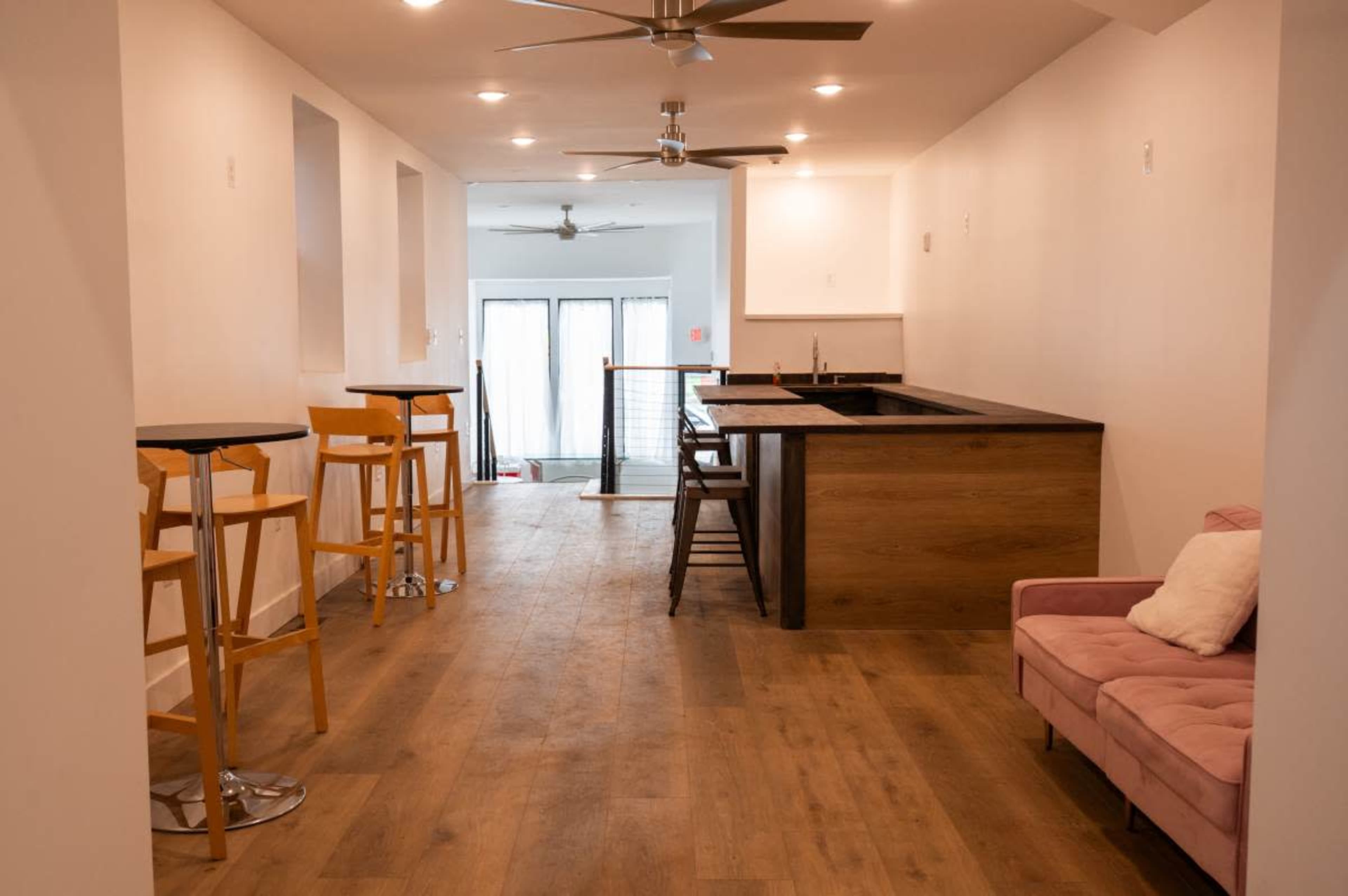The image shows a spacious interior with wooden flooring, a kitchenette featuring bar stools, and a pink couch against the wall.