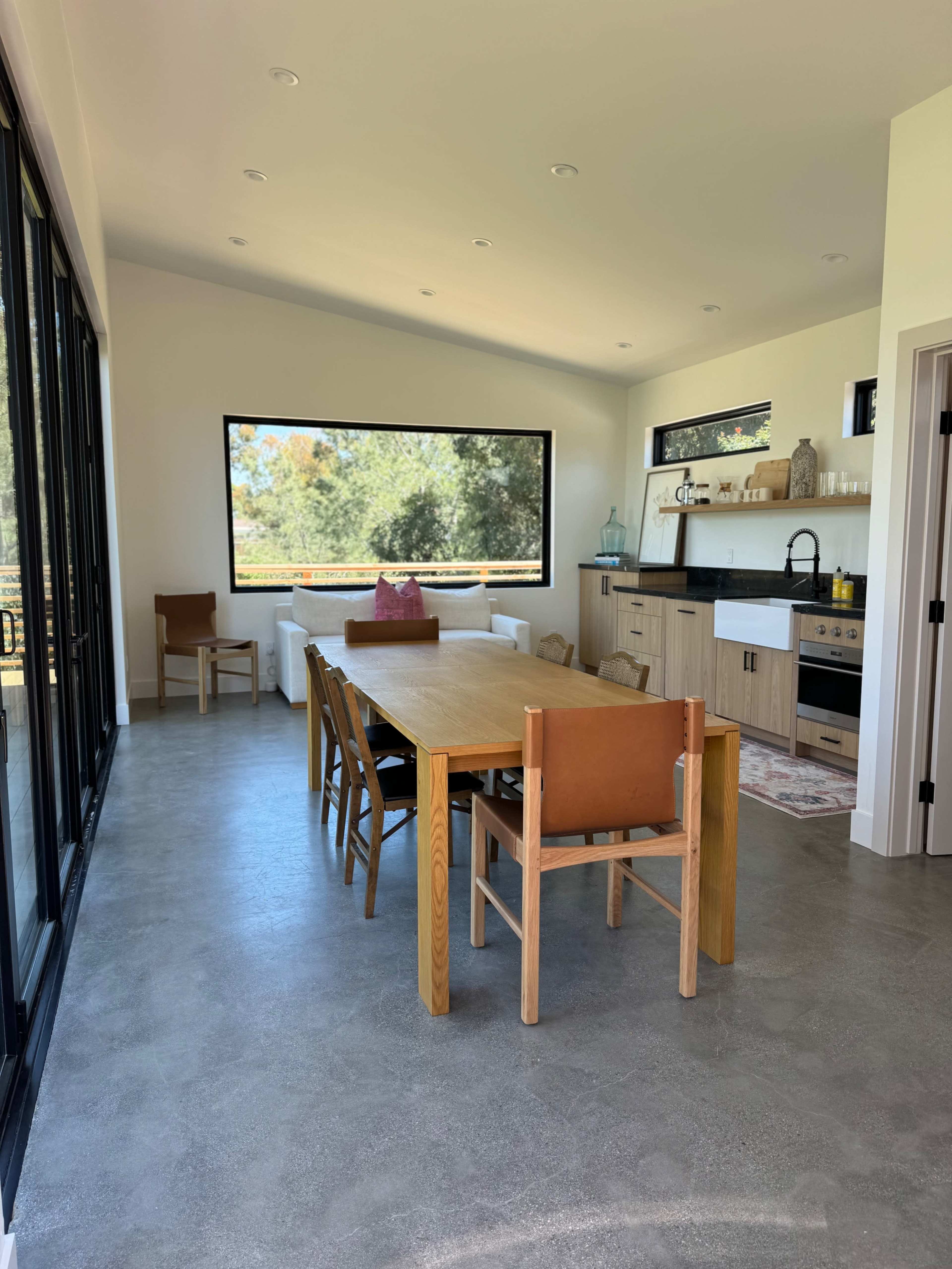 A bright, minimalist dining area with a large wooden table surrounded by chairs, and a kitchen area in the background featuring countertop appliances.