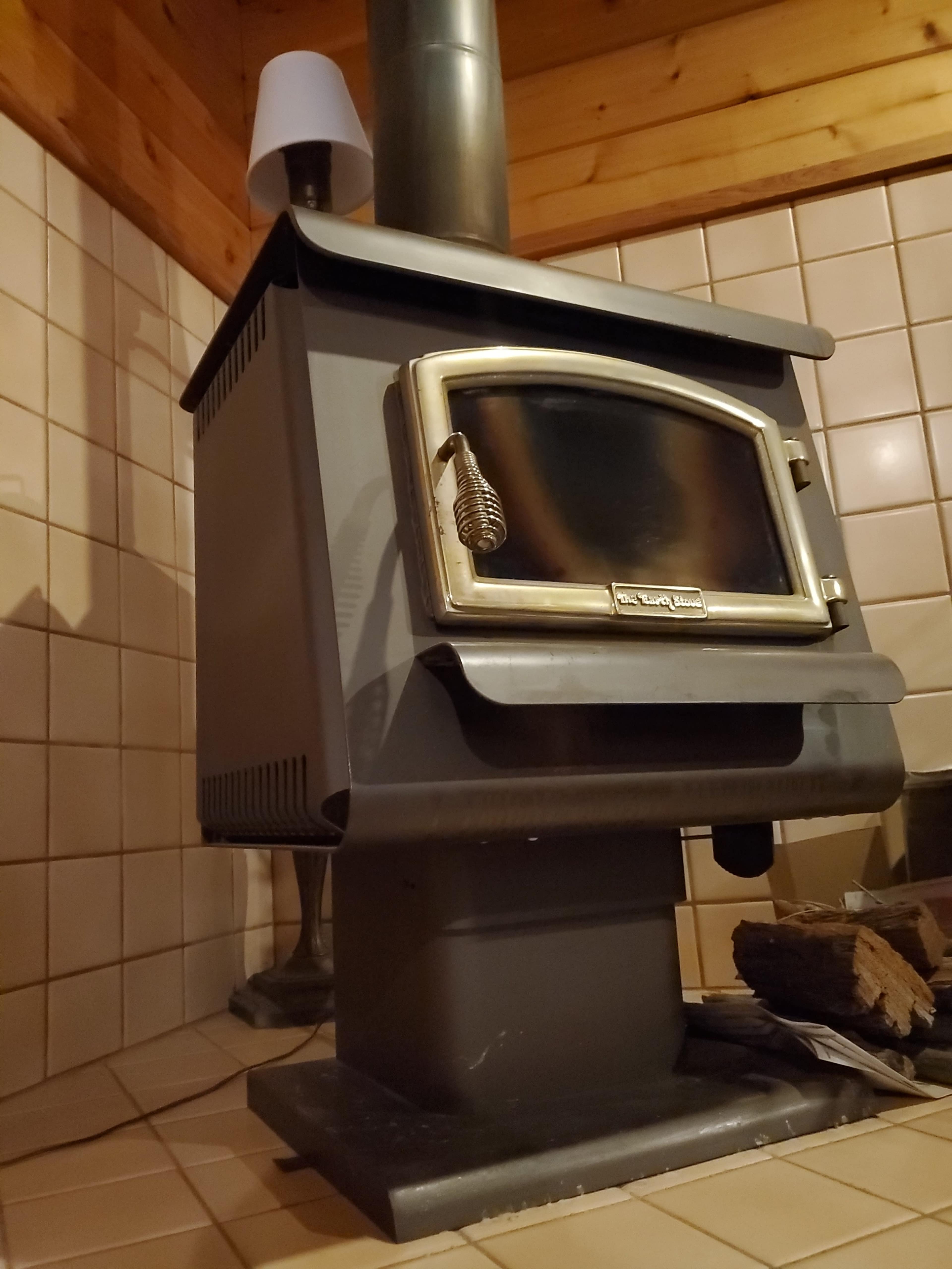 A gray wood-burning stove positioned in a corner of a tiled room, with a lamp nearby and logs stacked beside it.