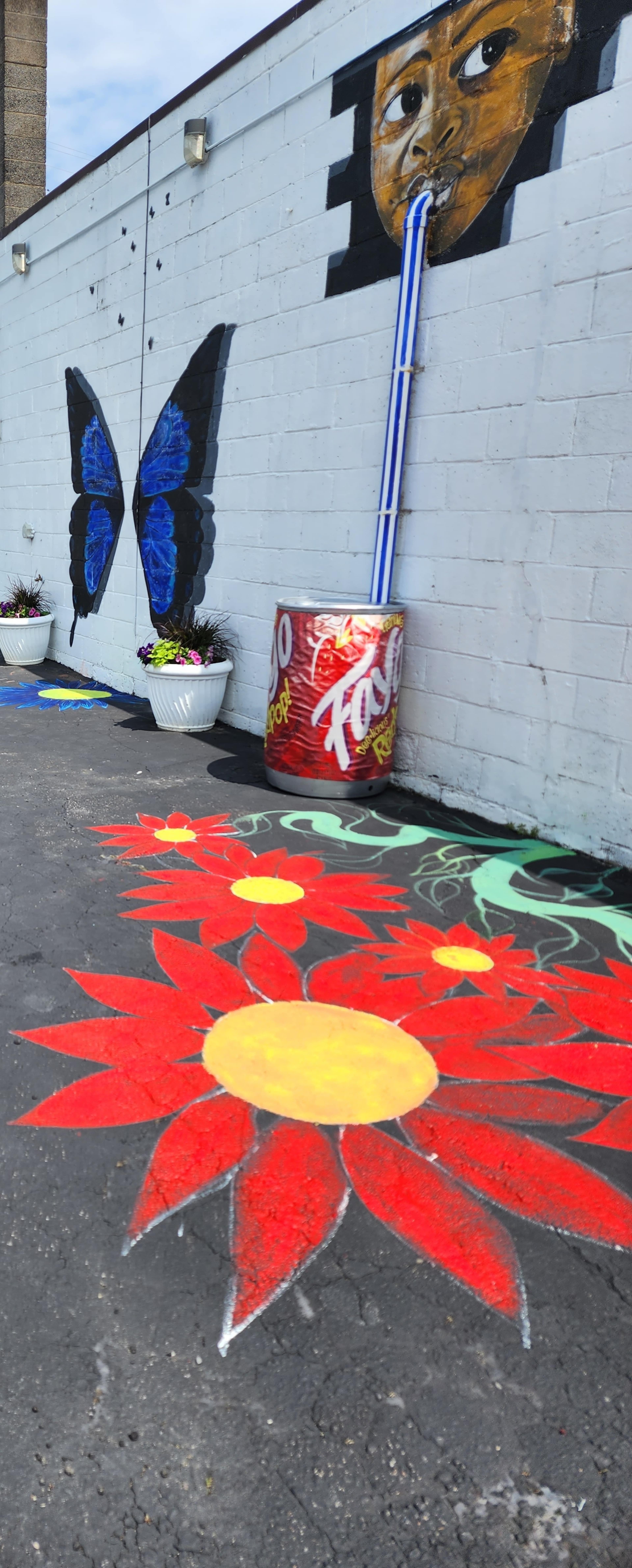The image shows a colorful mural on a wall featuring a butterfly, alongside a painted floor with large red flowers and a yellow center, near decorated pots.
