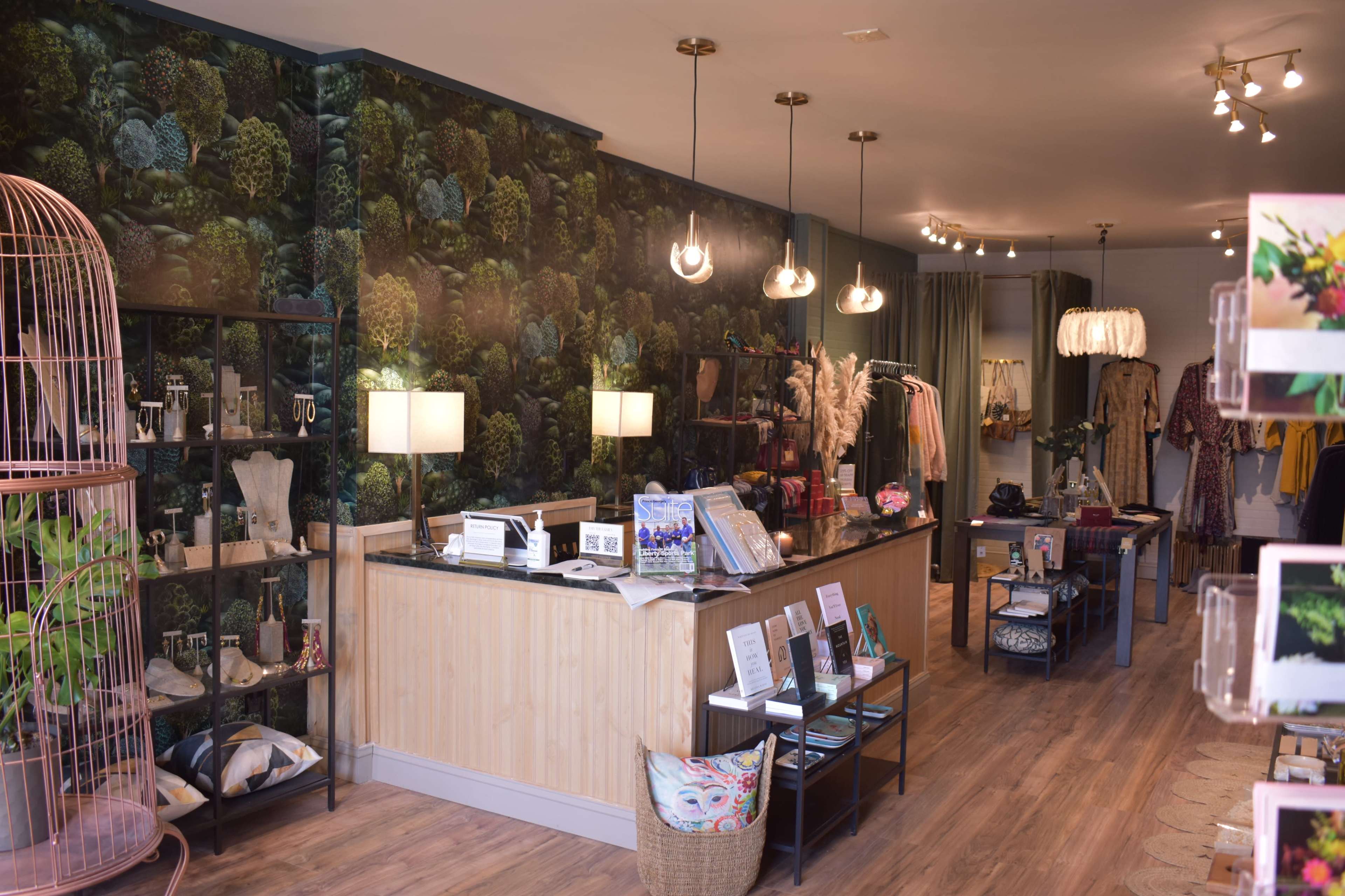 A retail store interior featuring a wooden counter, decorative wall wallpaper, and a variety of clothing and accessories displayed on racks and shelves.