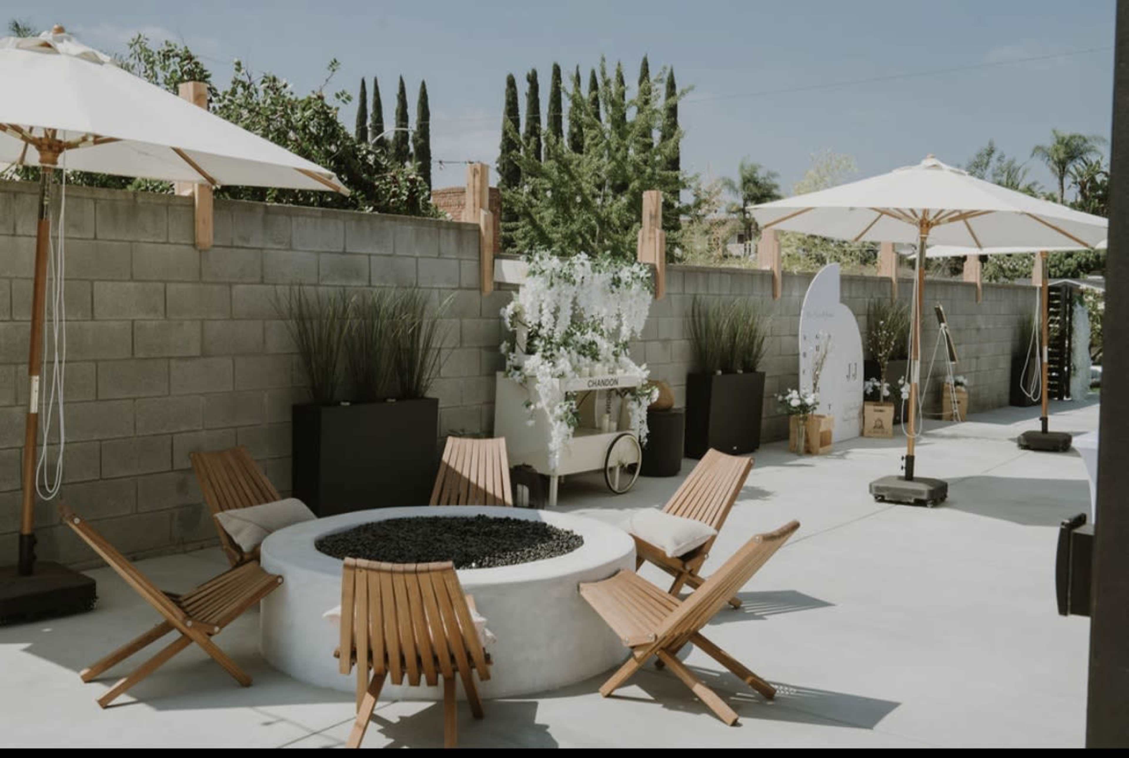 The image shows a modern outdoor space featuring a circular fire pit surrounded by wooden lounge chairs, two white umbrellas, and greenery along a gray wall.