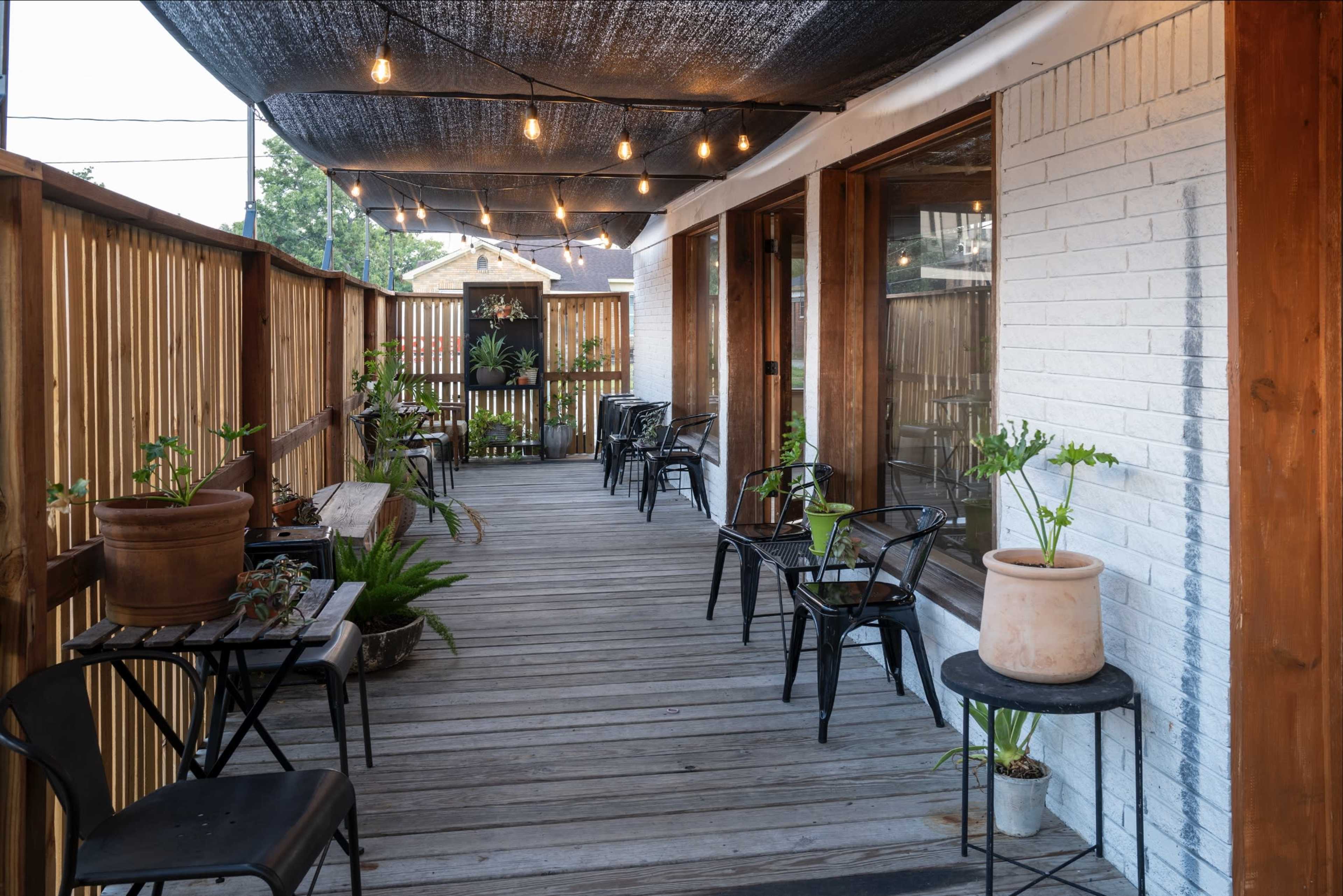 The image shows a wooden deck with potted plants and empty chairs under decorative string lights and a canopy.