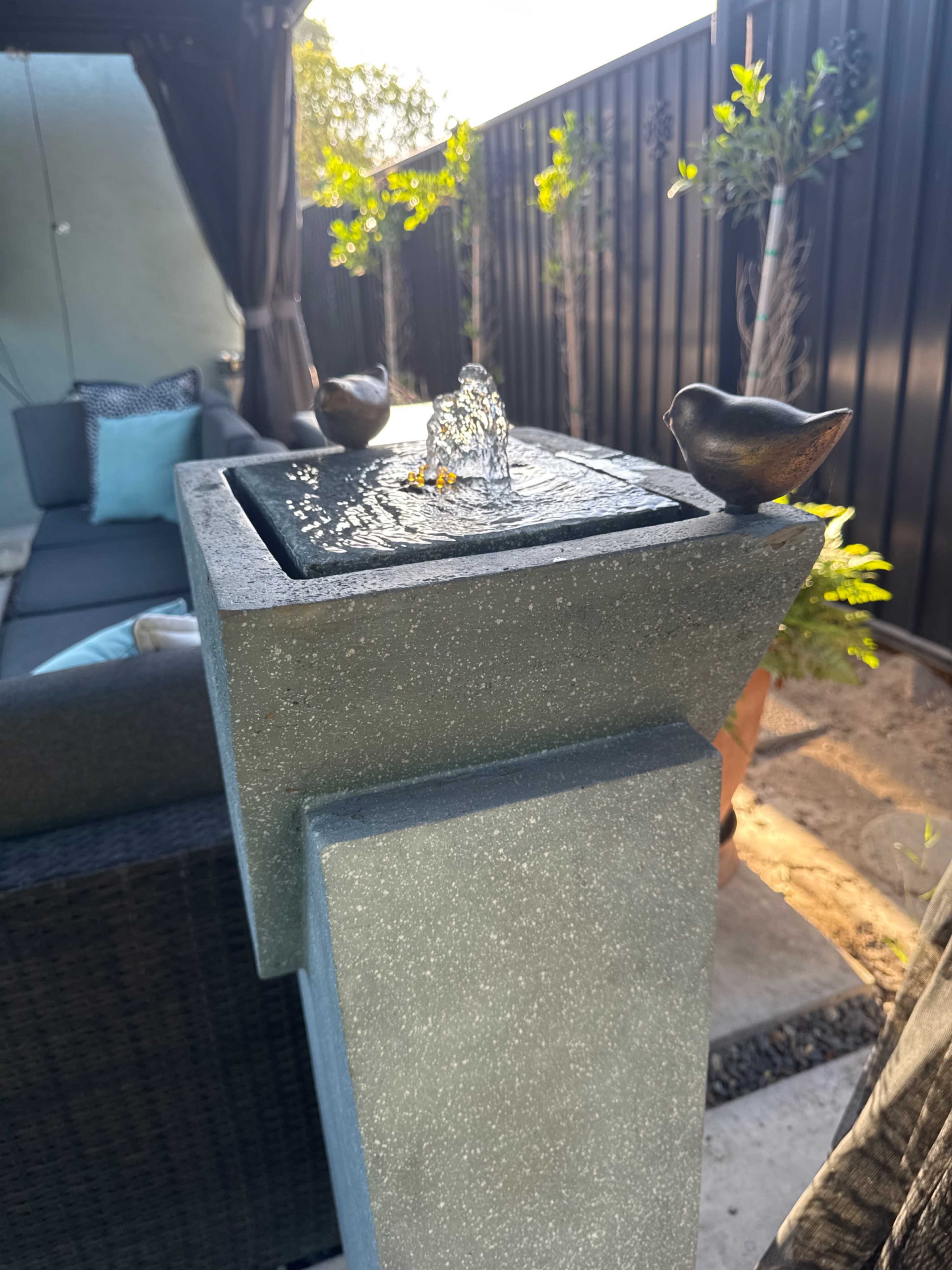 A small decorative fountain with two bird figures sits on a pedestal in a backyard setting.