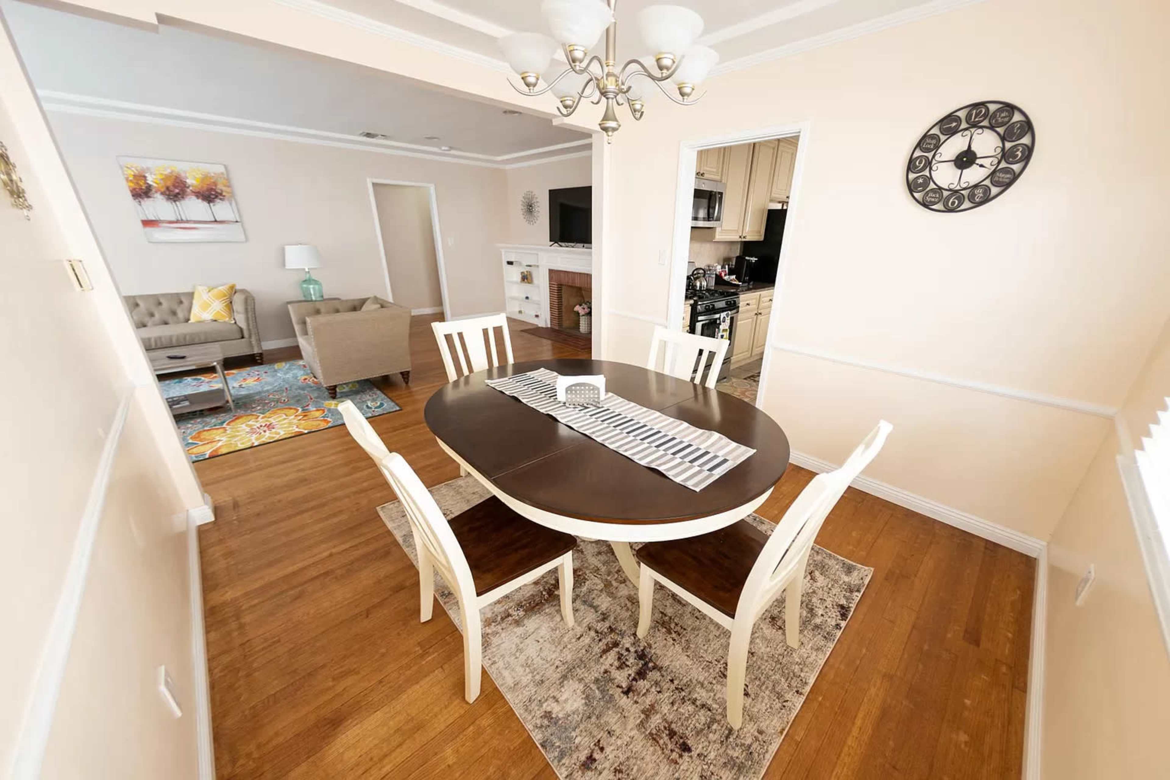 A dining area features an oval table surrounded by four chairs, with a decorative rug beneath, and an open view into a living room and kitchen.
