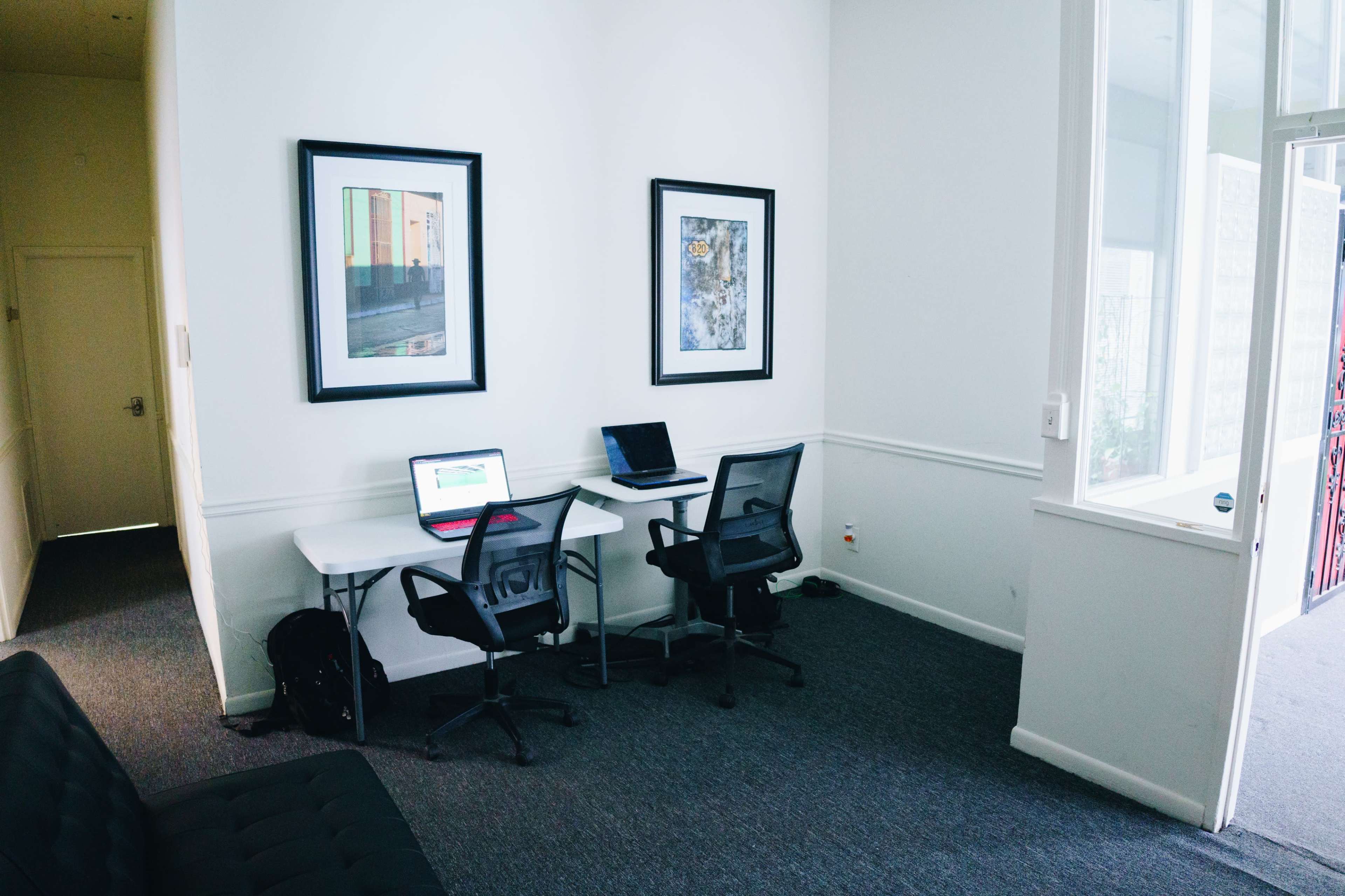 A small office space features two chairs and desks, each with a laptop, adjacent to a hallway and adorned with framed artwork on the walls.