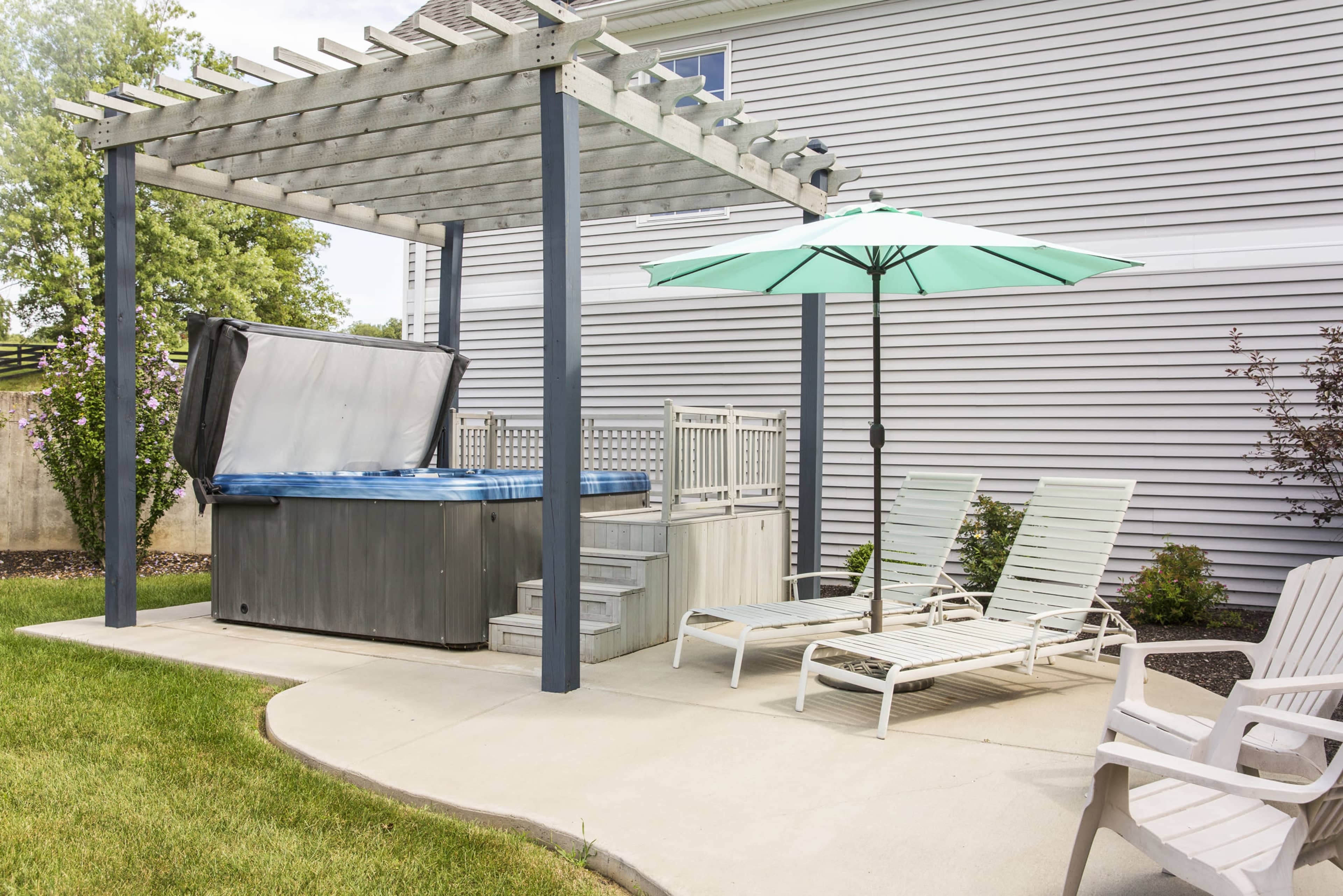 A hot tub is placed under a wooden pergola beside a patio featuring a green umbrella and lounge chairs.