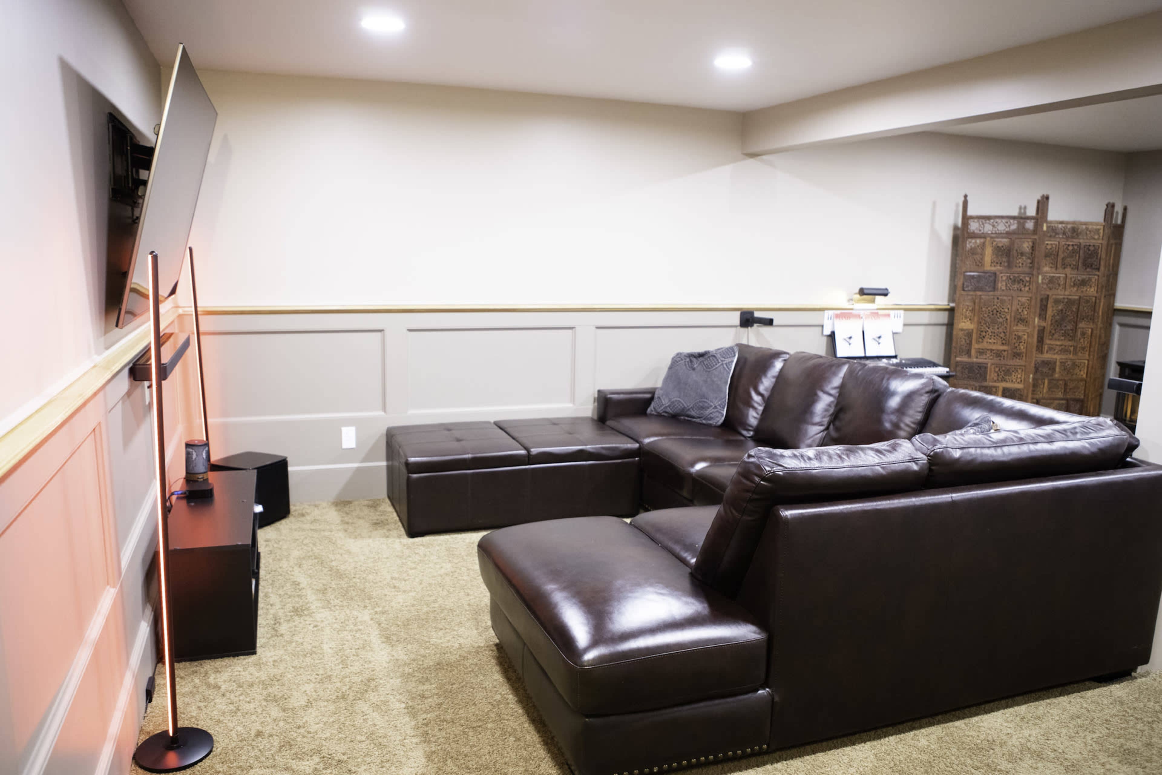 The image shows a cozy basement living room featuring a dark brown sectional sofa, a television mounted on the wall, and a light-colored carpet.