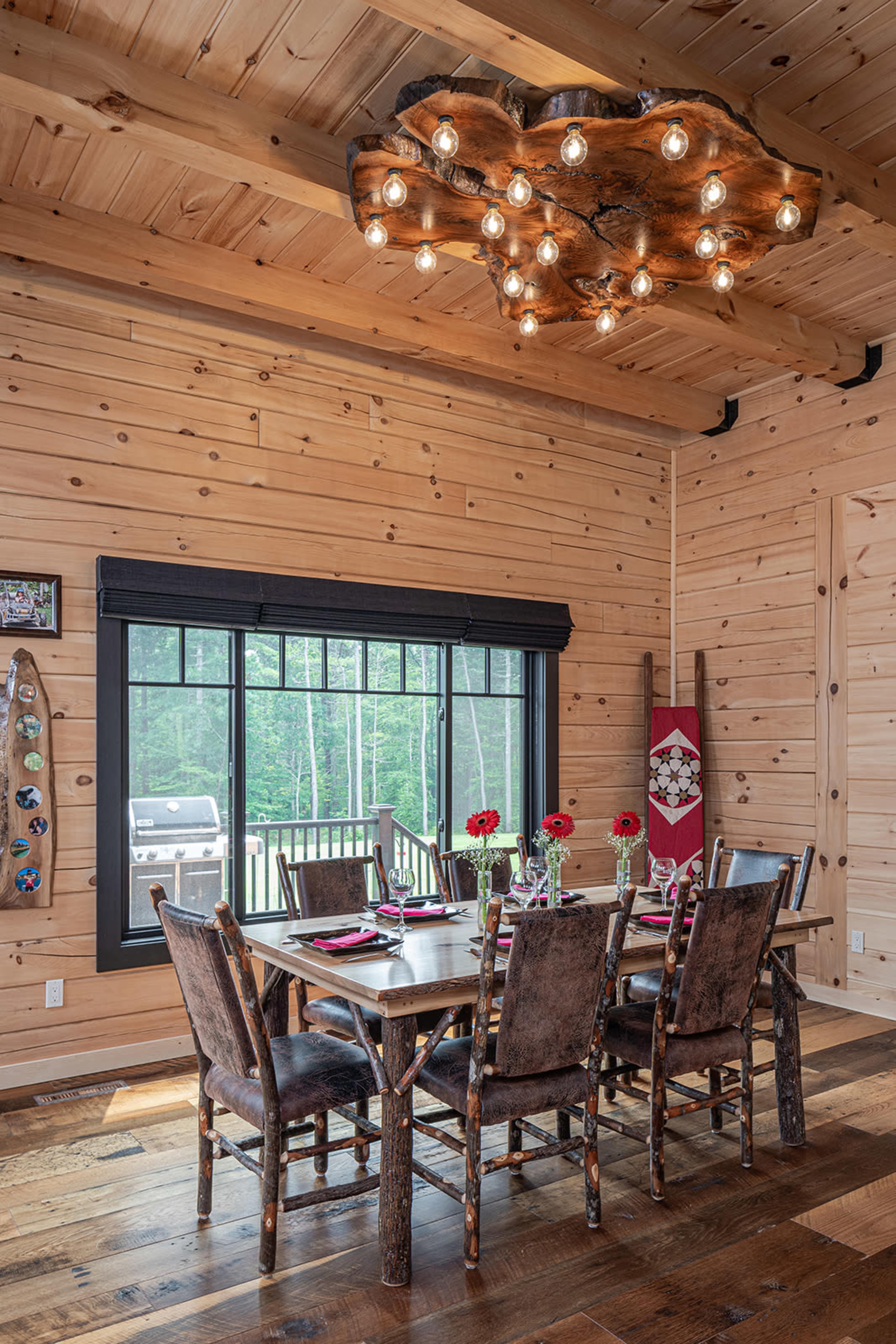 A rustic dining room features a large wooden table surrounded by chairs, with a chandelier made of wood and light bulbs hung overhead, and large windows letting in natural light.