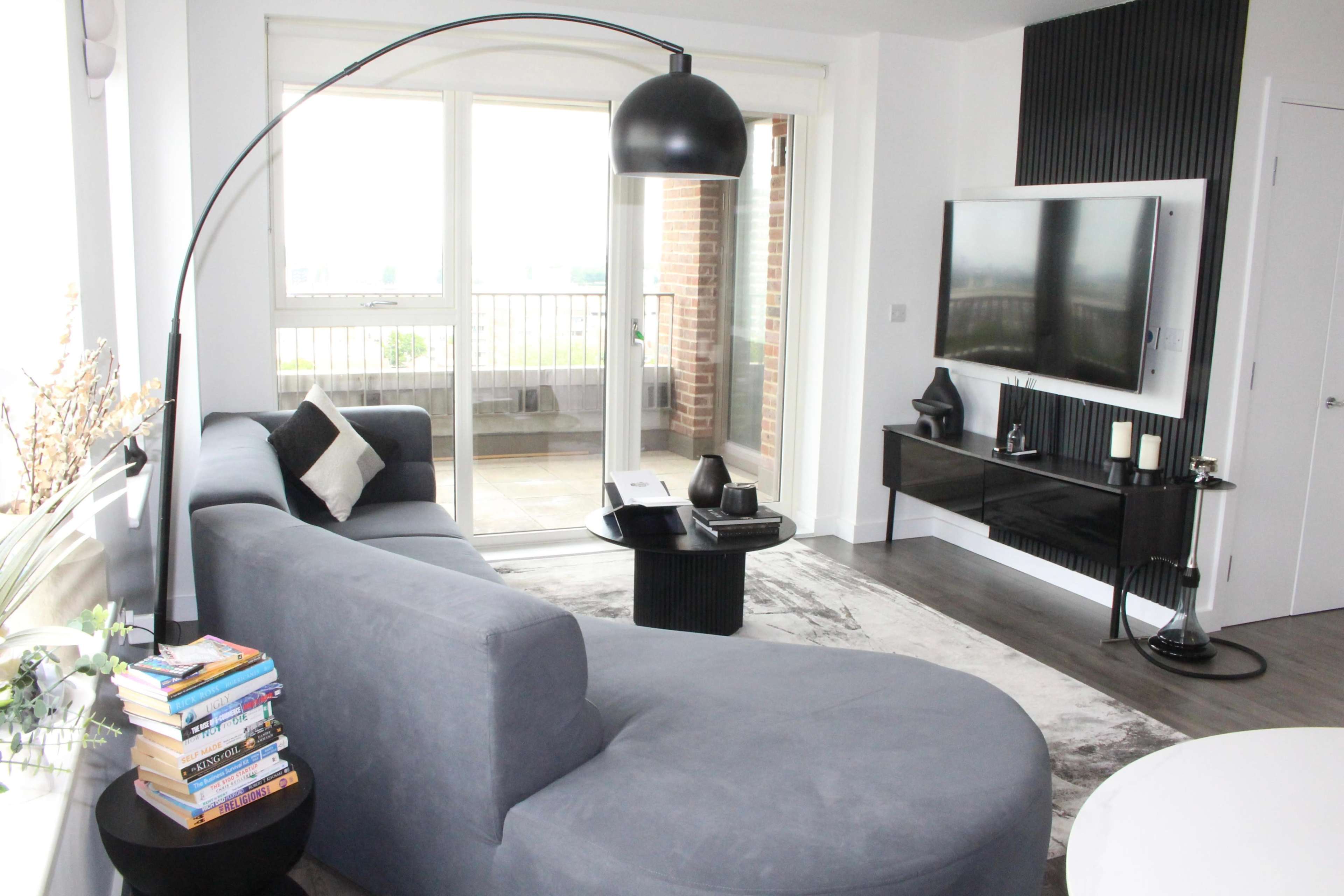 The image shows a modern living room featuring a curved gray sofa, a floor lamp, a black television unit, and a balcony visible through large sliding glass doors.