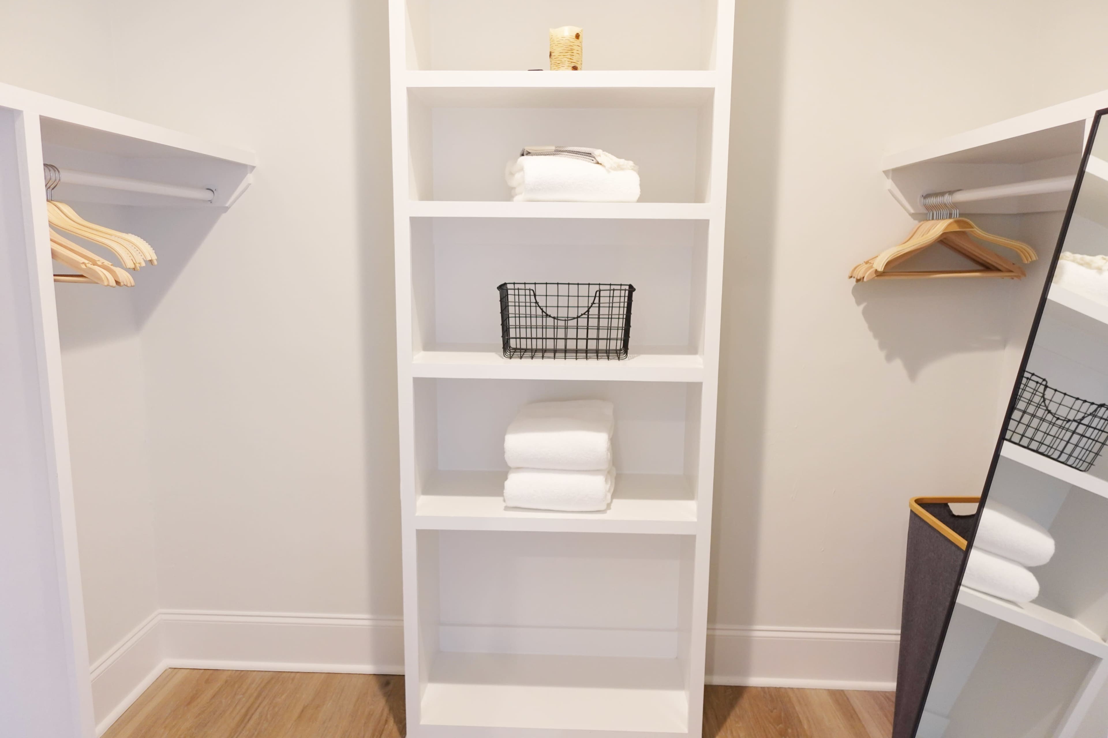 A minimalist closet features a white shelving unit with neatly stacked towels, a wire basket, and wooden hangers on either side.