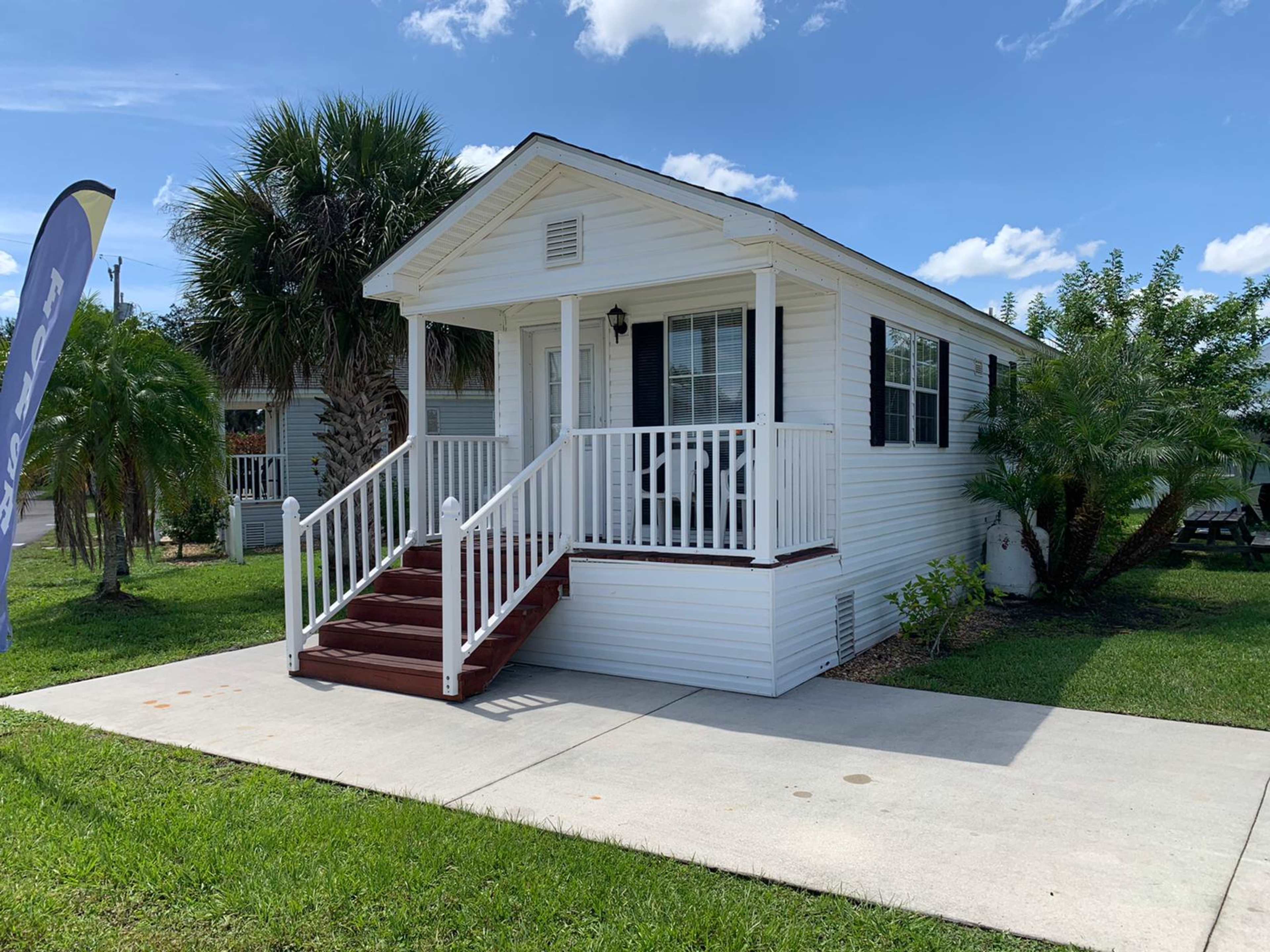 The image shows a white, single-story house with a porch and steps, situated on a grassy lawn with palm trees in the background.