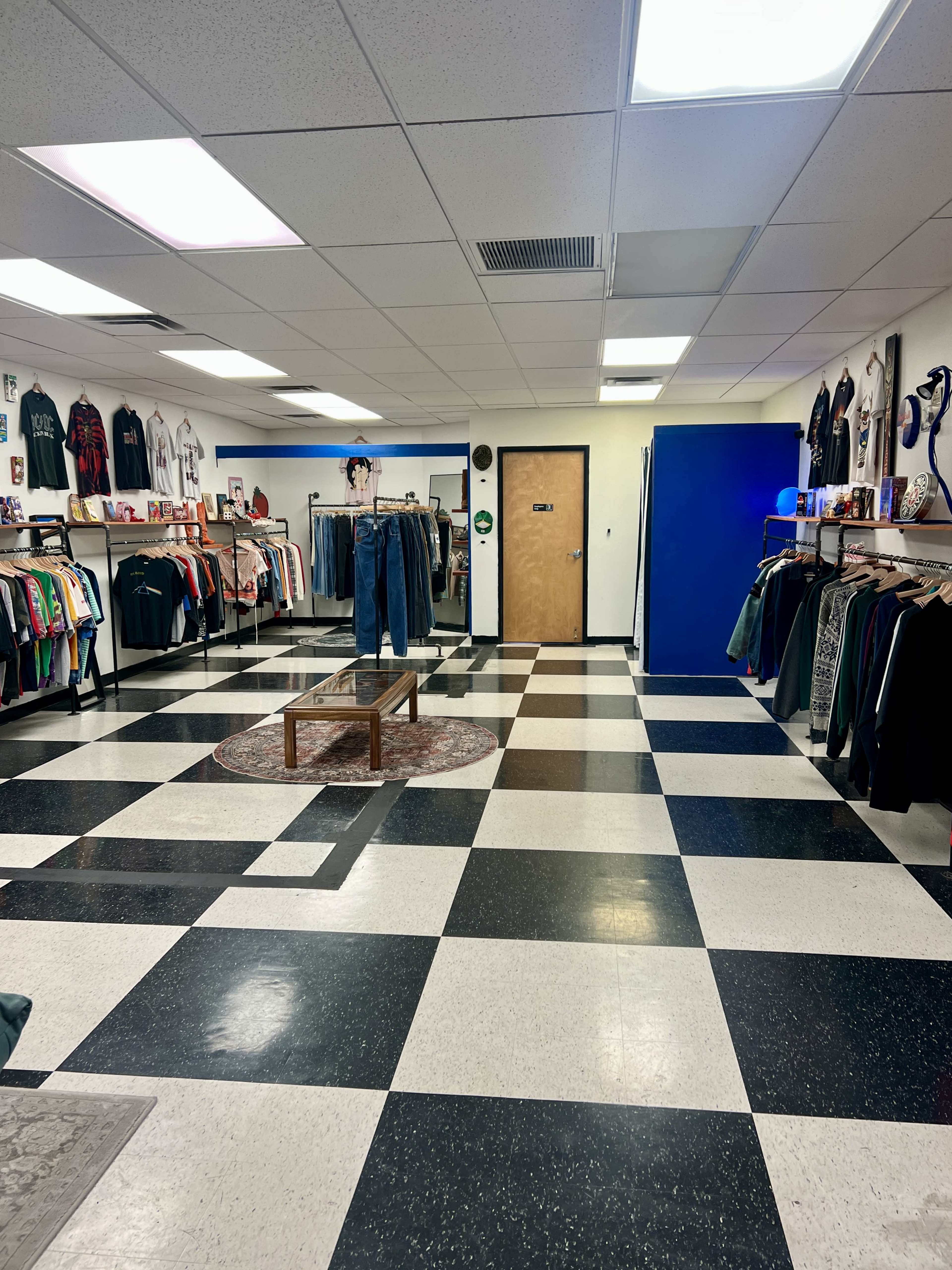 The image shows the interior of a clothing store with black and white checkered flooring, clothing racks lining the walls, and a central table.