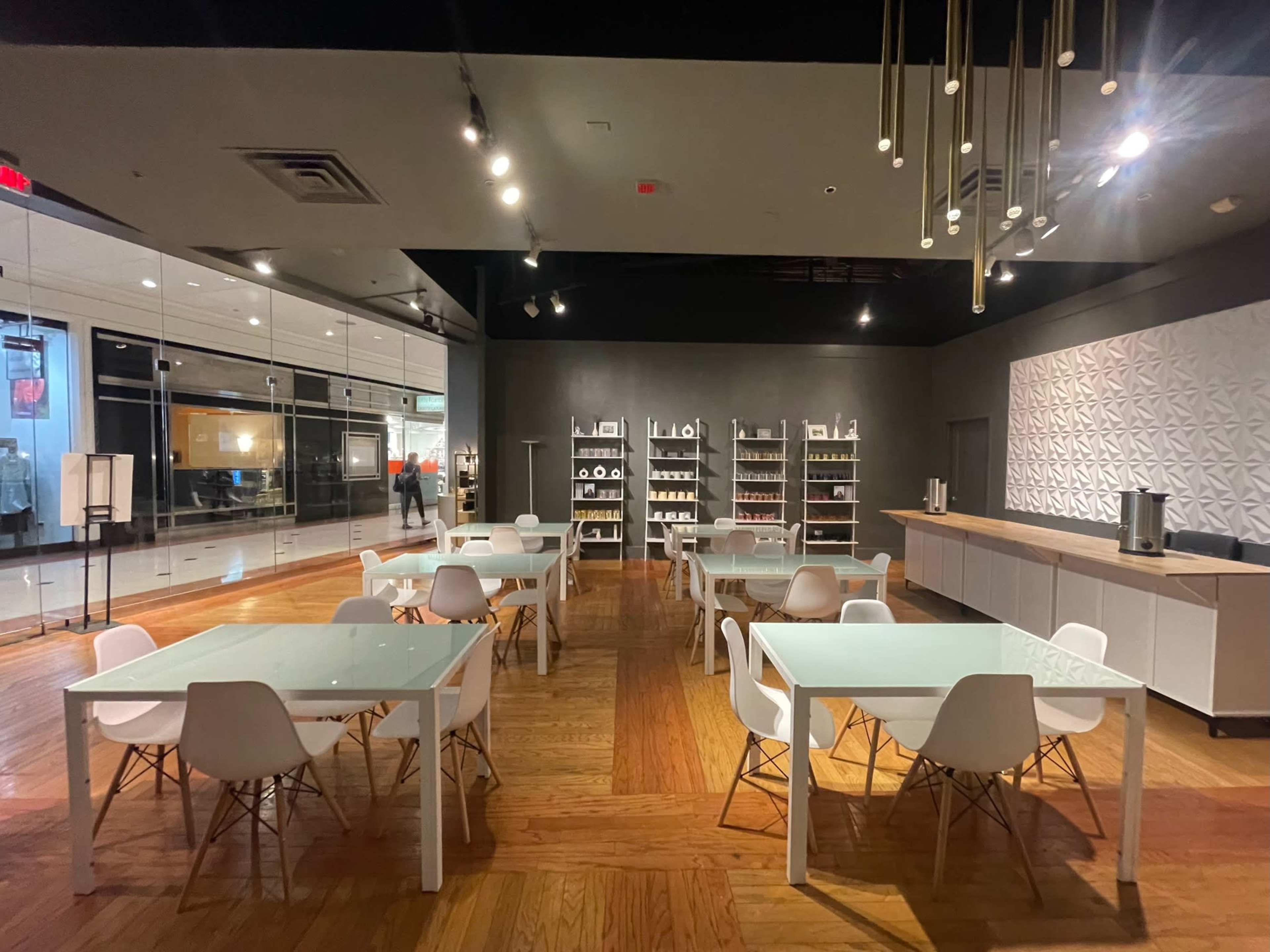 A modern, spacious room with several white tables and chairs arranged neatly, alongside shelves displaying various white dishware.