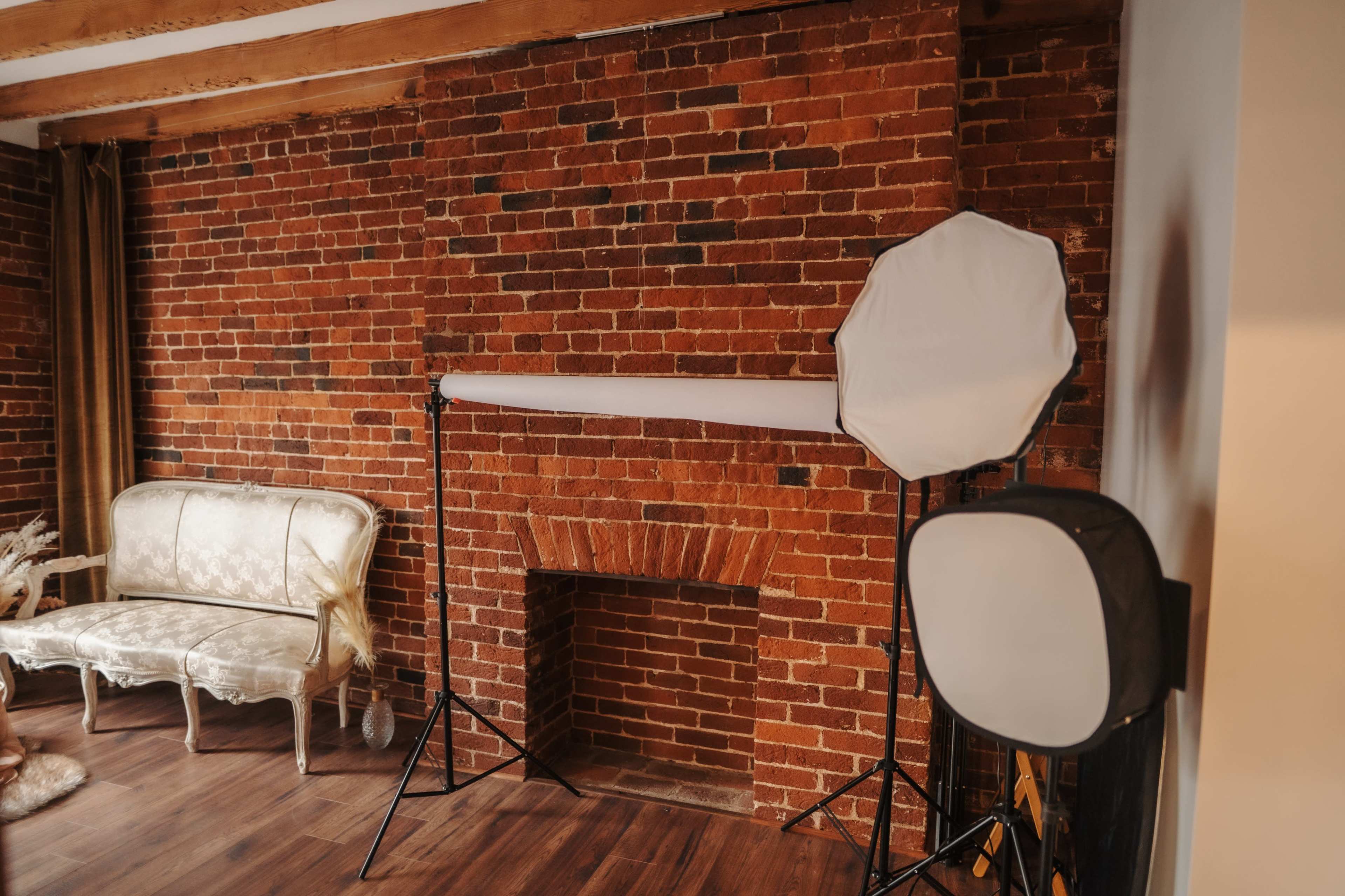 The image shows a room with a brick wall, a vintage-style sofa, and photography lighting equipment set up near a fireplace.