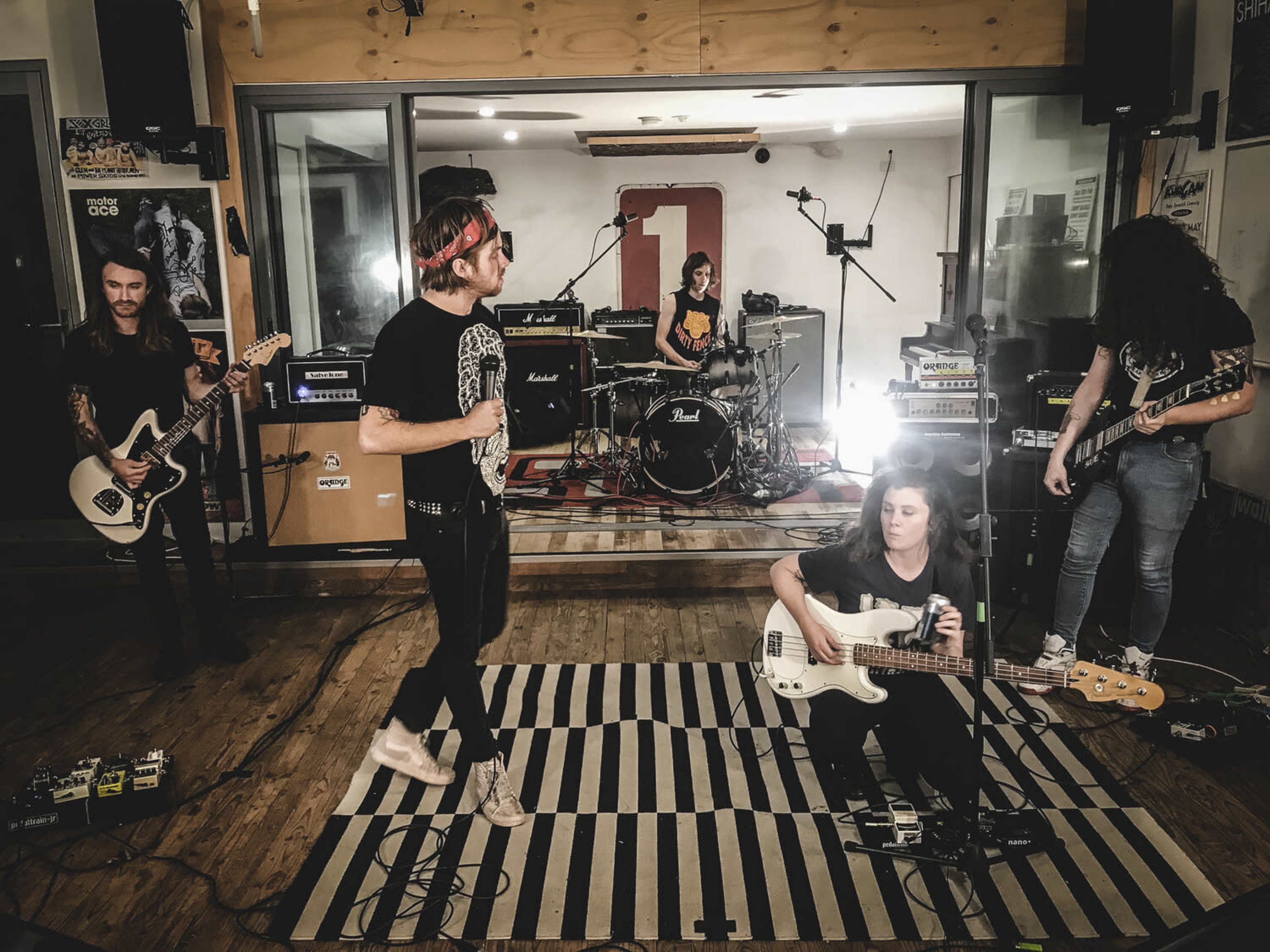 A five-member band performs in a studio with amplifiers and instruments arranged around them.