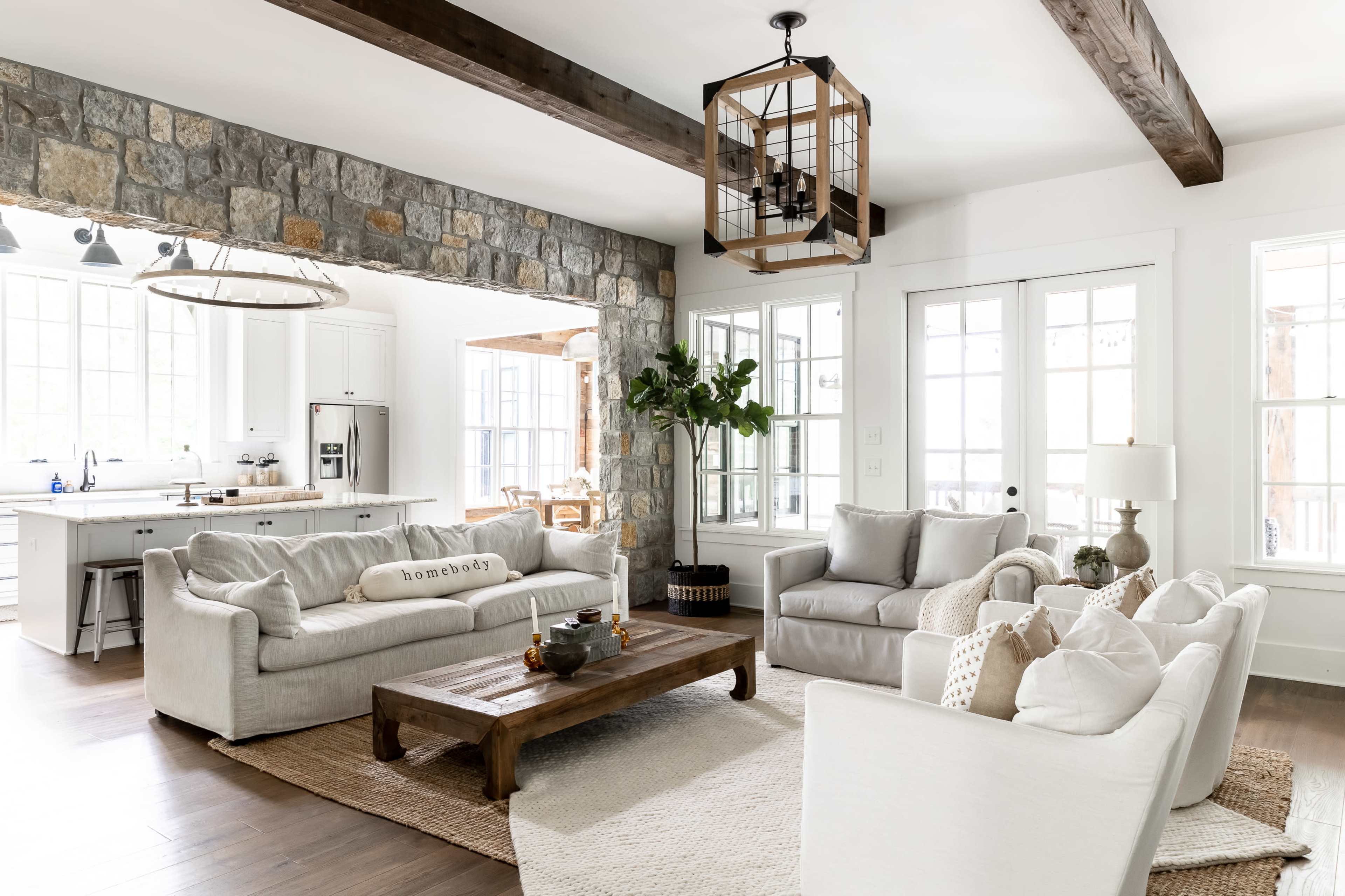A modern living room with two sofas facing a wooden coffee table, set against a stone wall, leading into a kitchen area with white cabinetry and large windows.