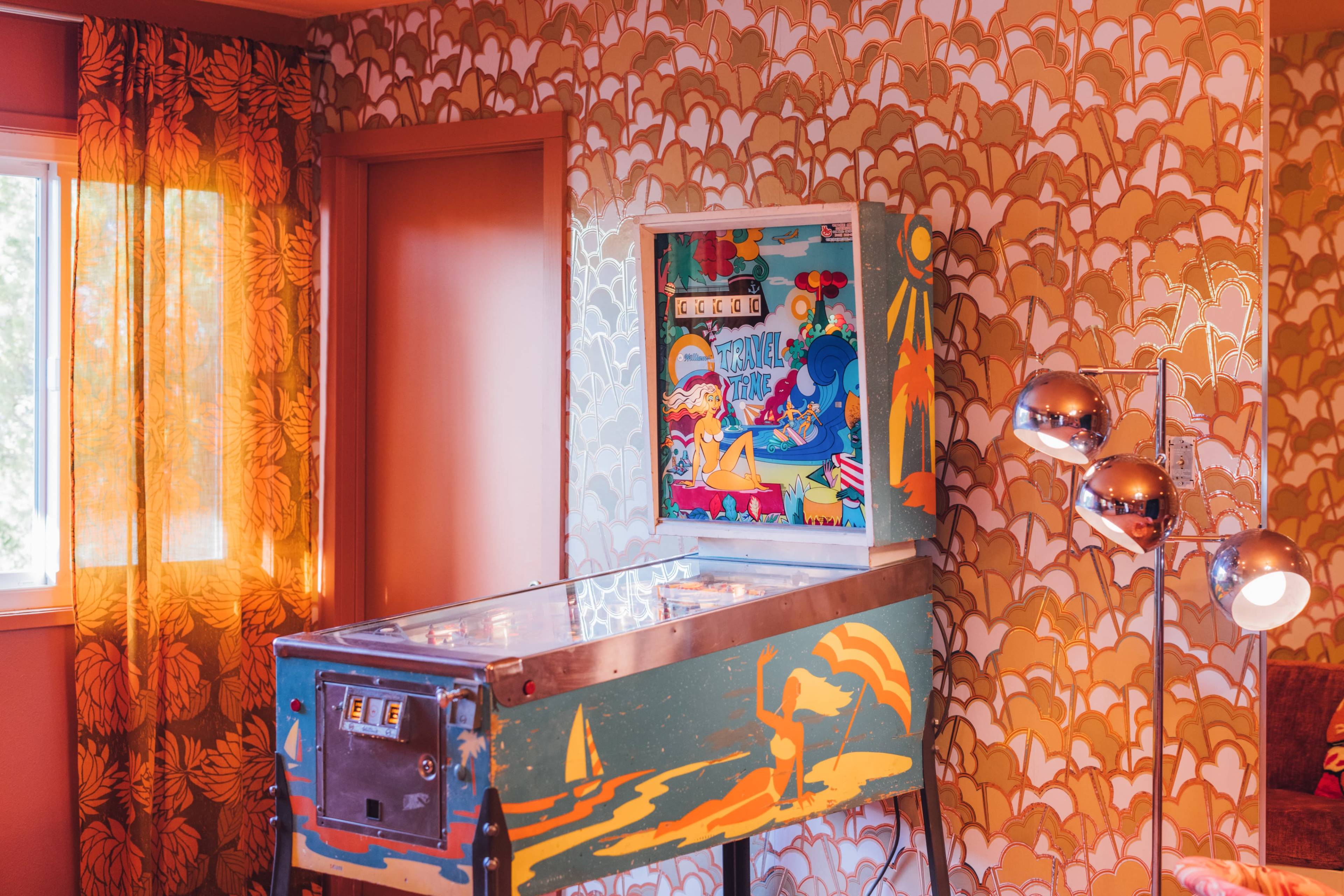 A vintage pinball machine stands against a patterned wall adorned with retro wallpaper in a brightly lit room.