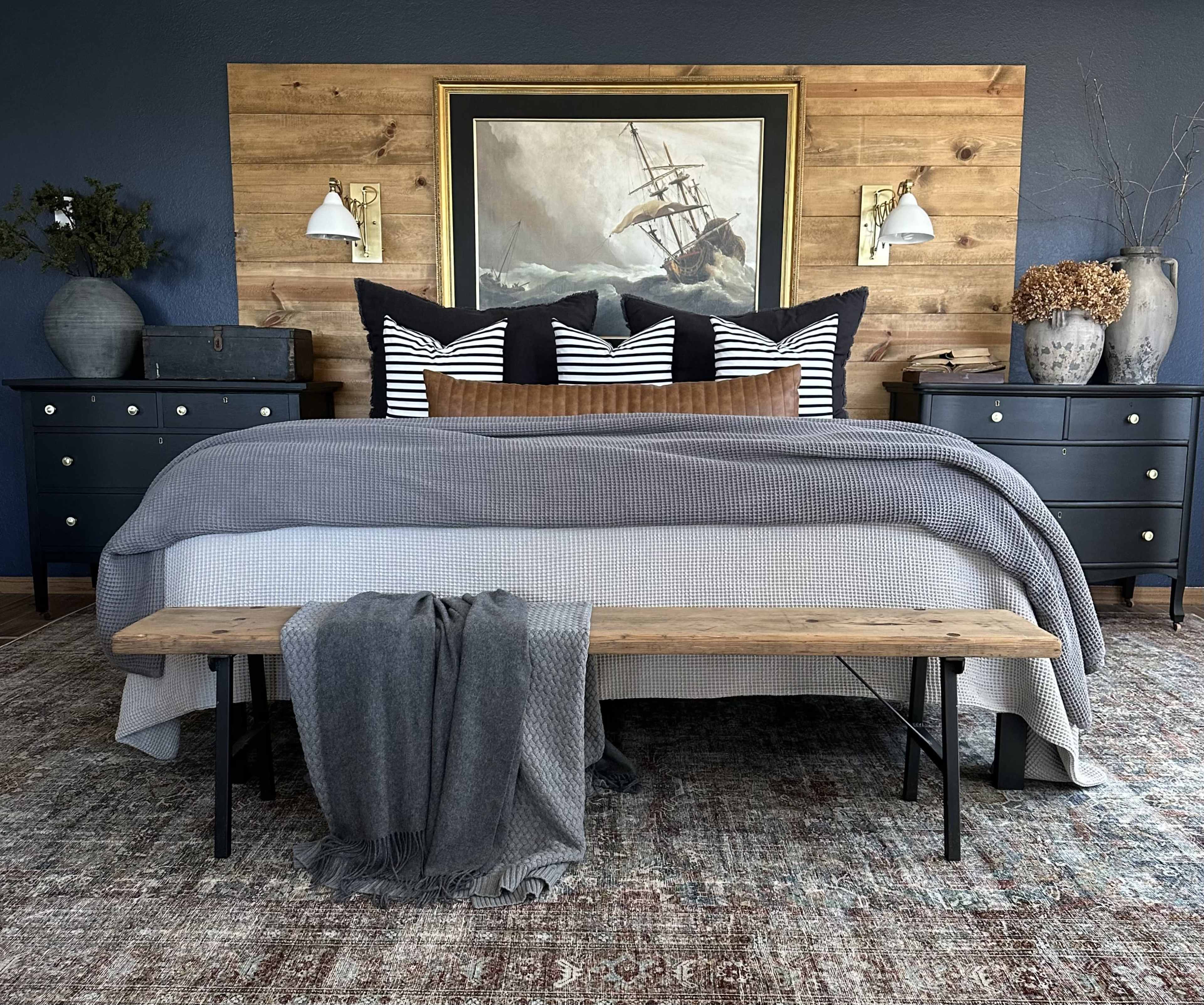 A neatly arranged bedroom featuring a large bed with a gray blanket, striped pillows, a wooden bench at the foot, and decorative elements against a wooden accent wall.