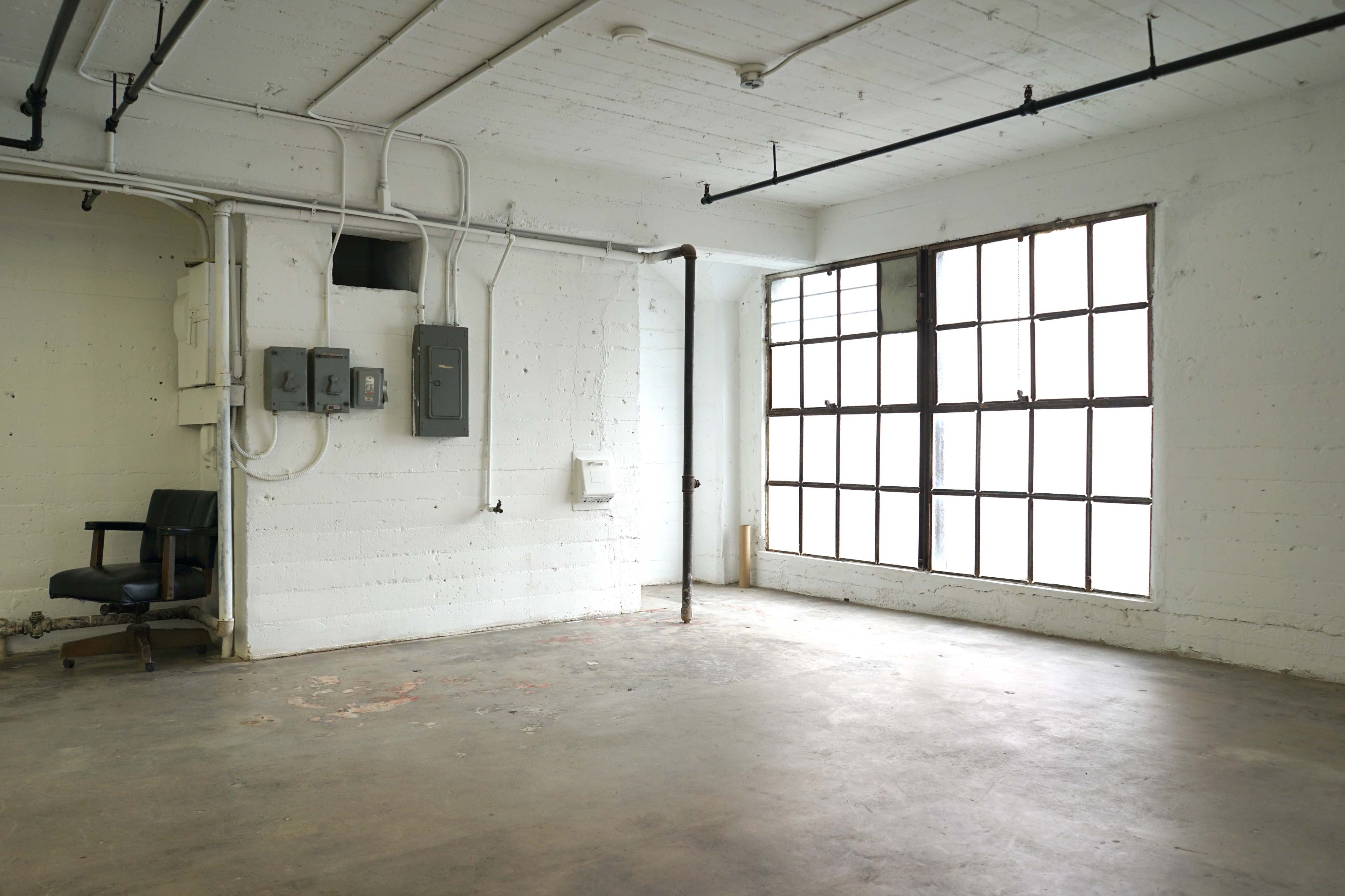 The image shows an empty industrial space with a concrete floor, exposed pipes, and a large window allowing natural light.