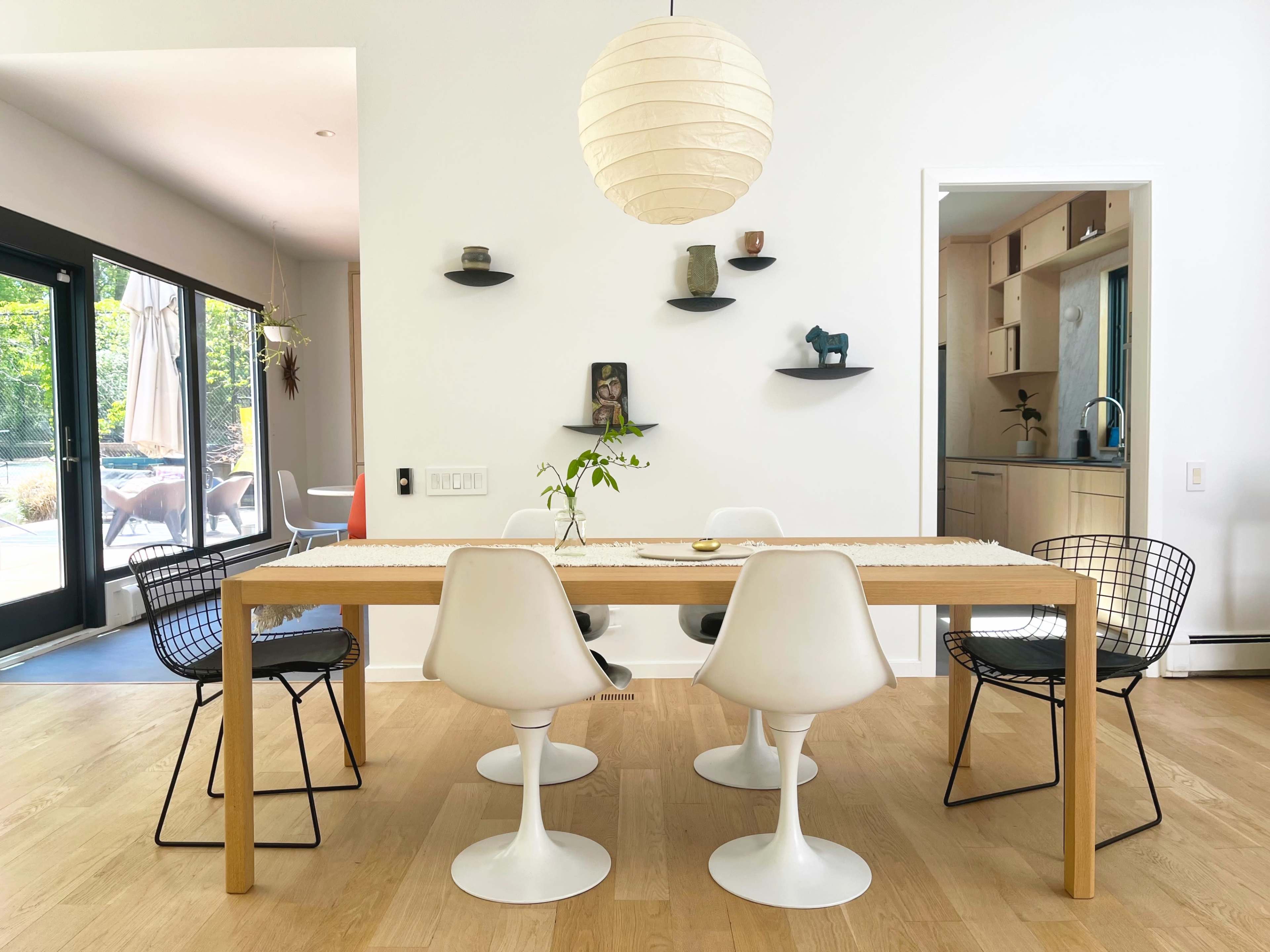 A modern dining area features a wooden table surrounded by white and black chairs, with a pendant light overhead and shelves displaying decorative items on the walls.
