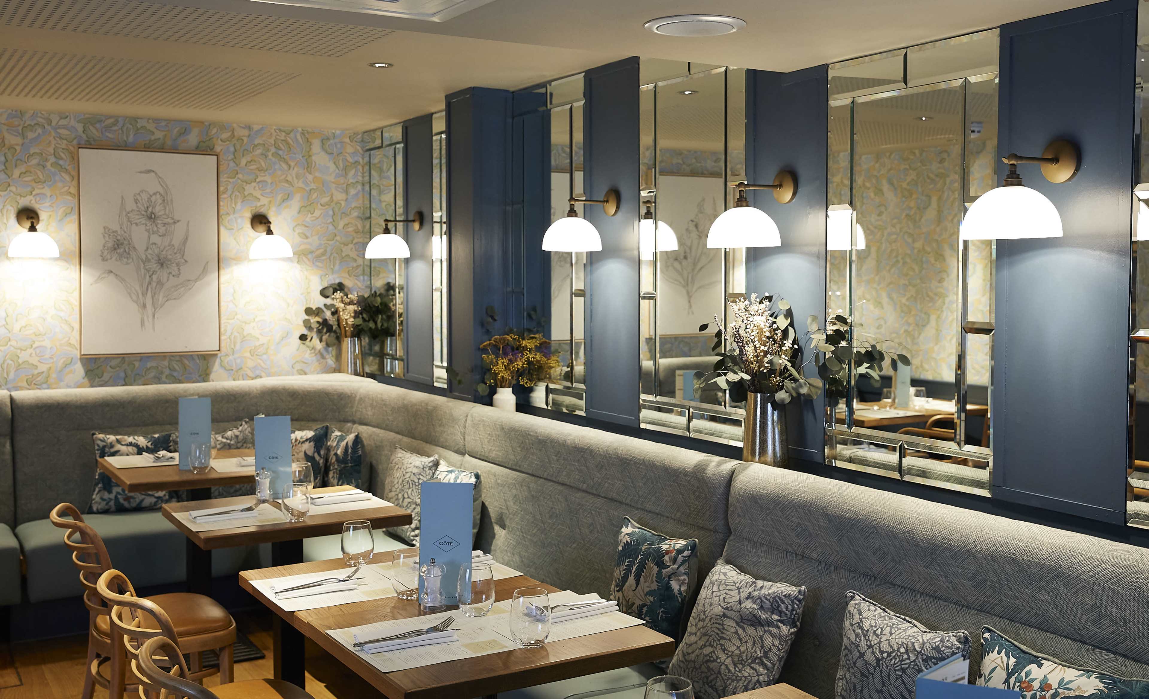 The image shows a stylish restaurant interior featuring blue floral wallpaper, wooden tables, and soft lighting from pendant lamps.