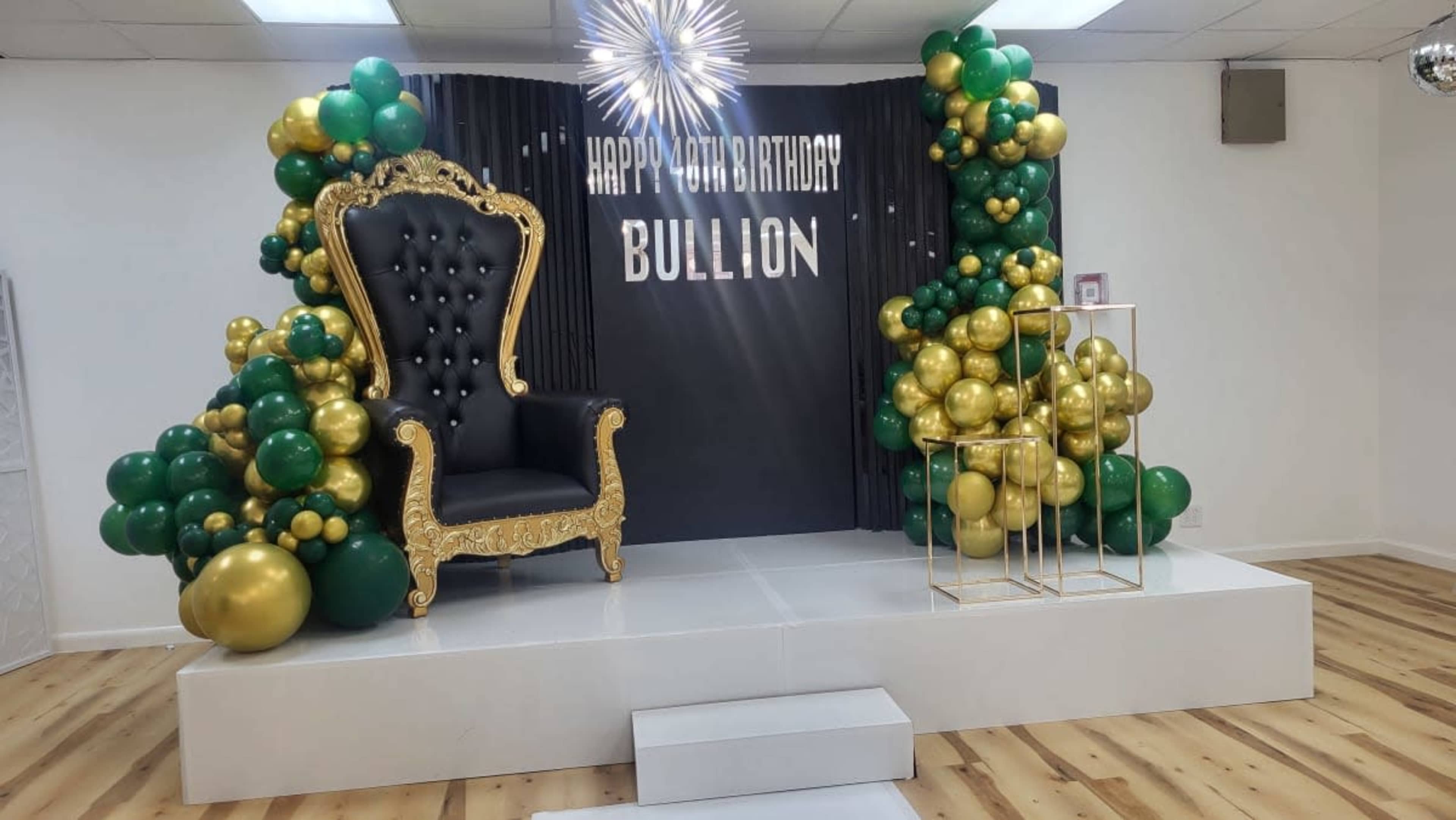 A decorated stage with a black and gold throne, a backdrop that reads "Happy 6th Birthday Bullion," and clusters of green and gold balloons on either side.