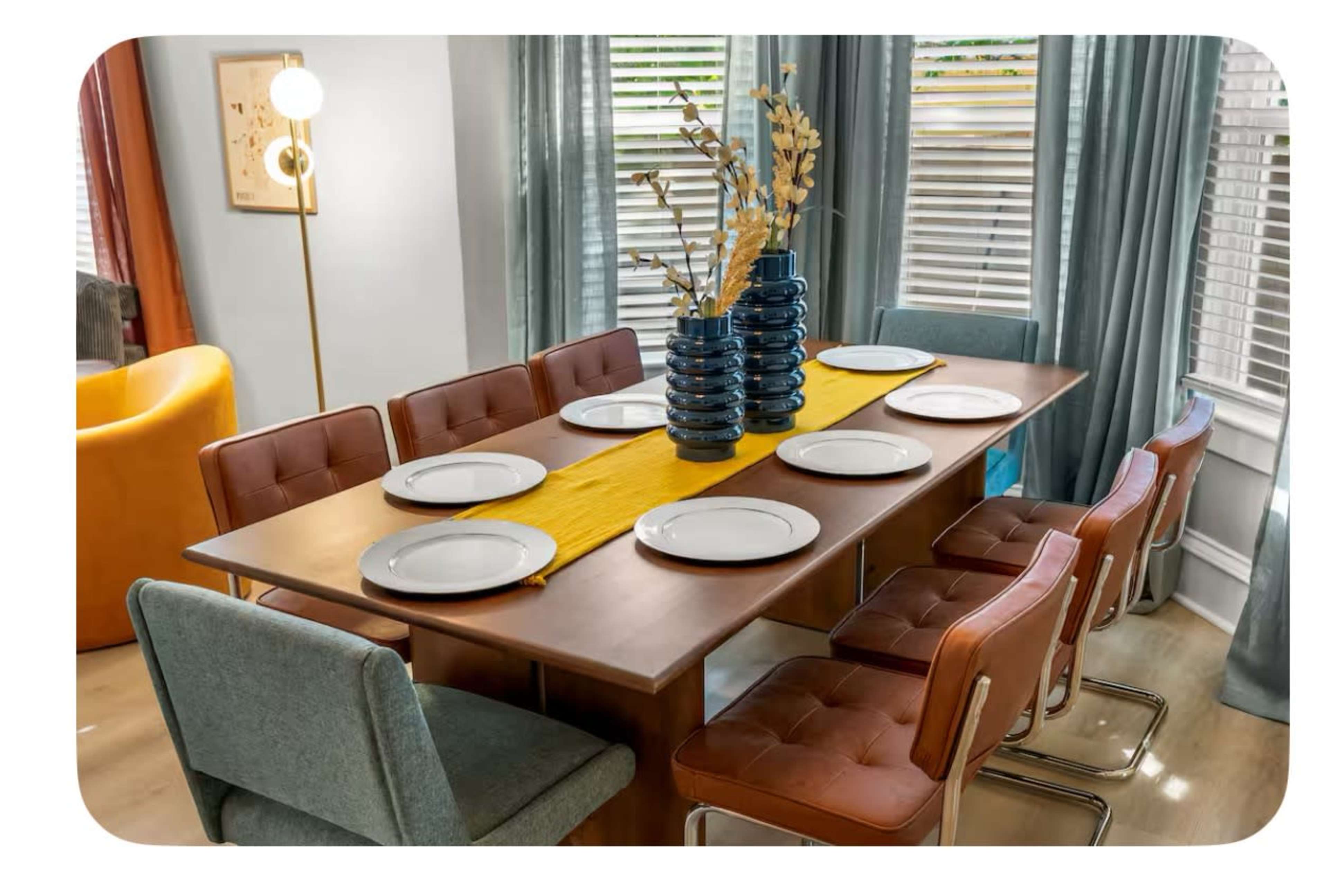 A modern dining room features a large wooden table set with eight plates, surrounded by a mix of brown and blue chairs, highlighted by a decorative vase and a yellow table runner.