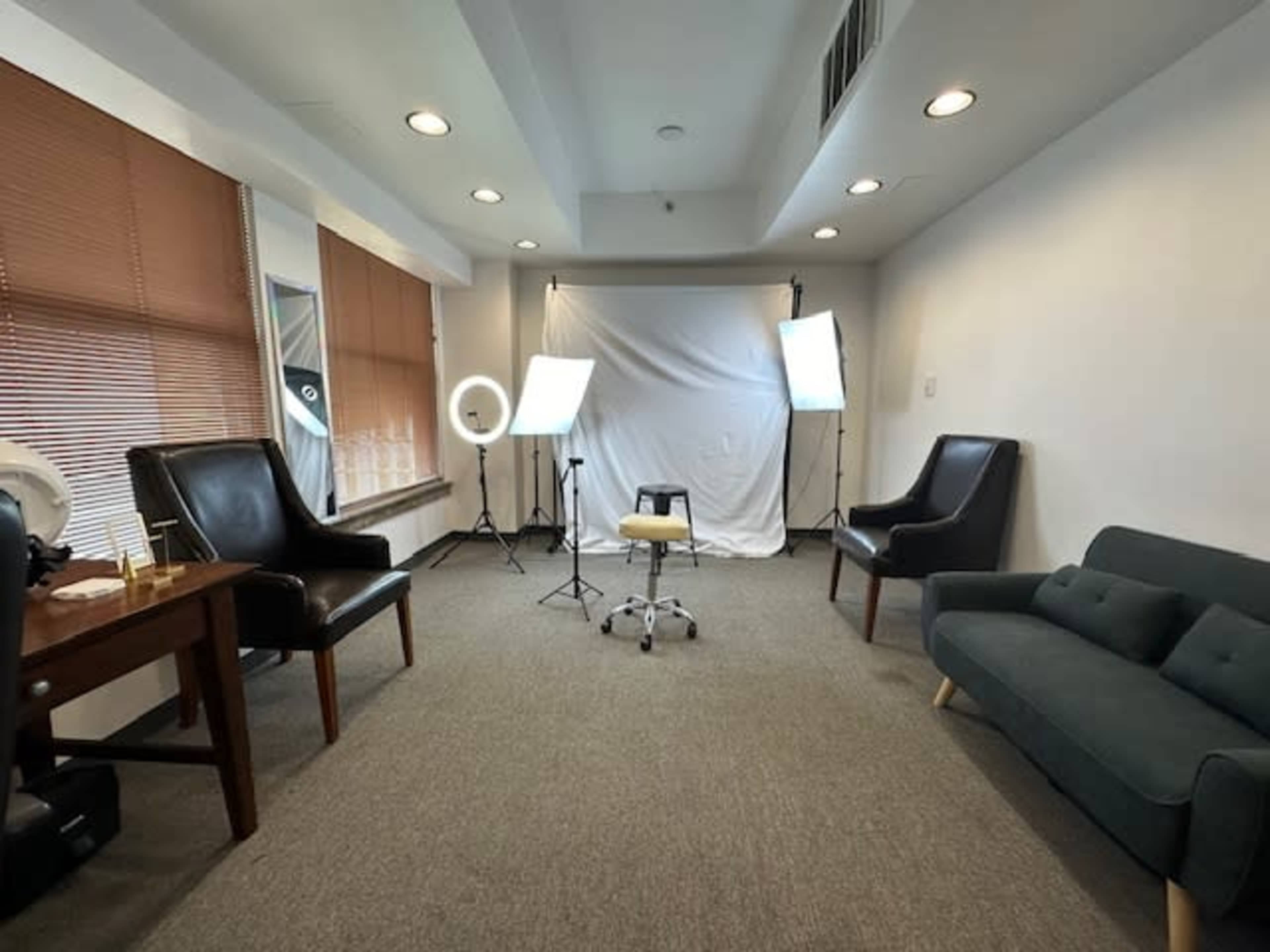 The image shows a well-lit photo studio setup featuring two studio lights, a backdrop, a chair in the center, and seating areas along the walls.