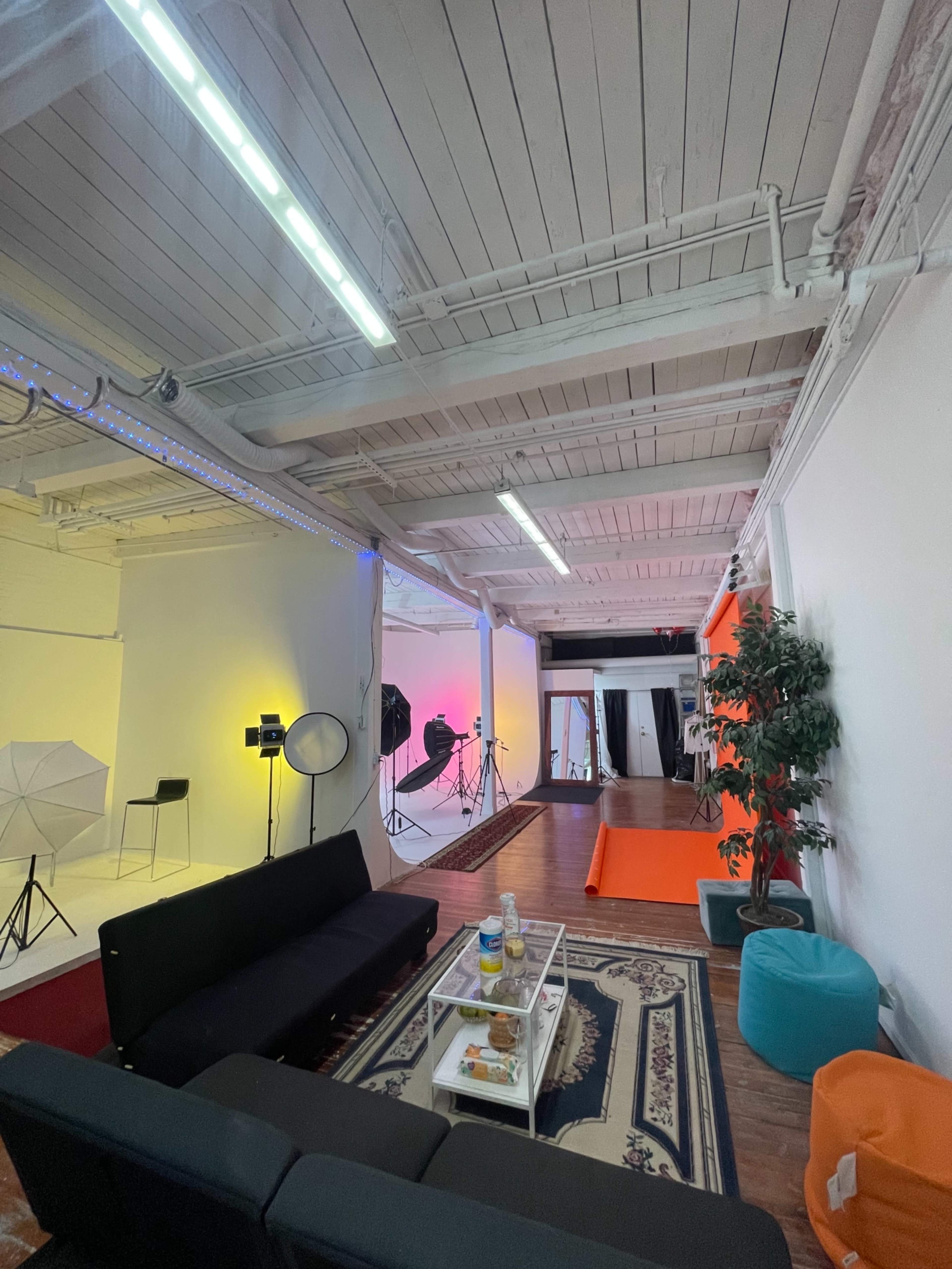 The image shows a spacious photography studio with white walls, professional lighting equipment, and a seating area featuring a black couch and colorful bean bags.