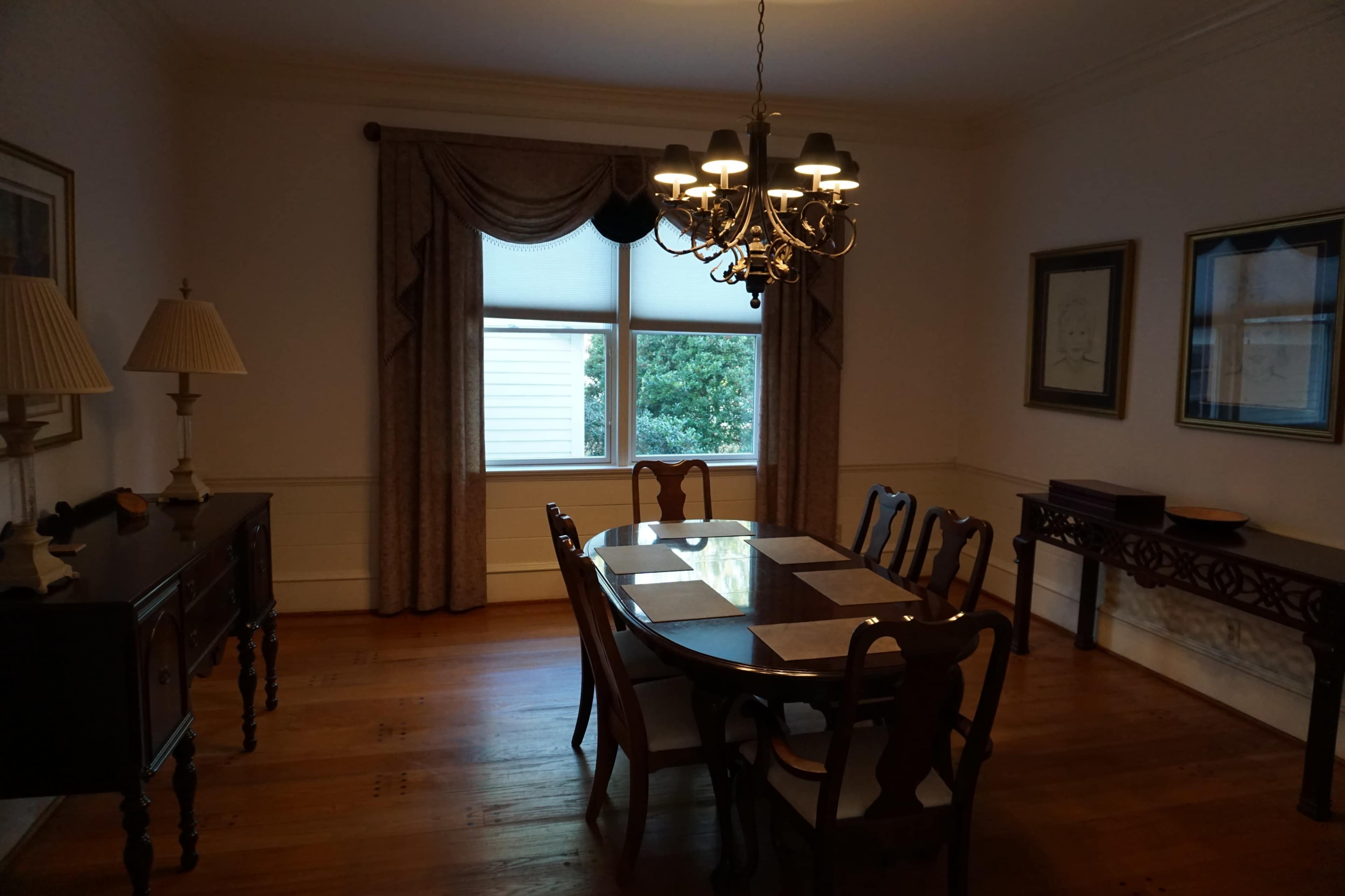 A dining room featuring a large wooden table surrounded by chairs, with a chandelier and curtains framing a window.