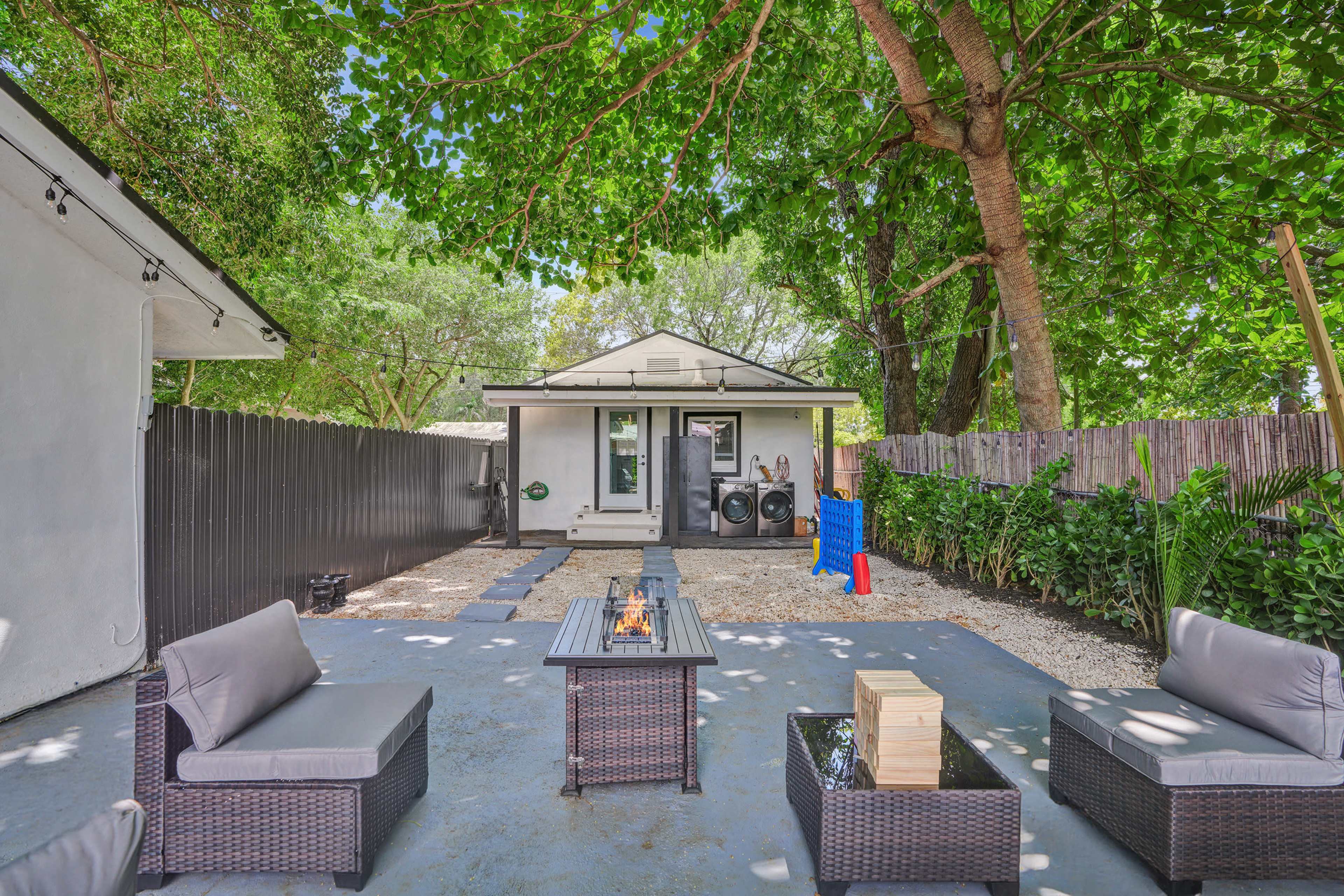 The image shows a backyard area with seating arrangements made of wicker furniture around a fire pit, bordered by greenery and a house in the background.