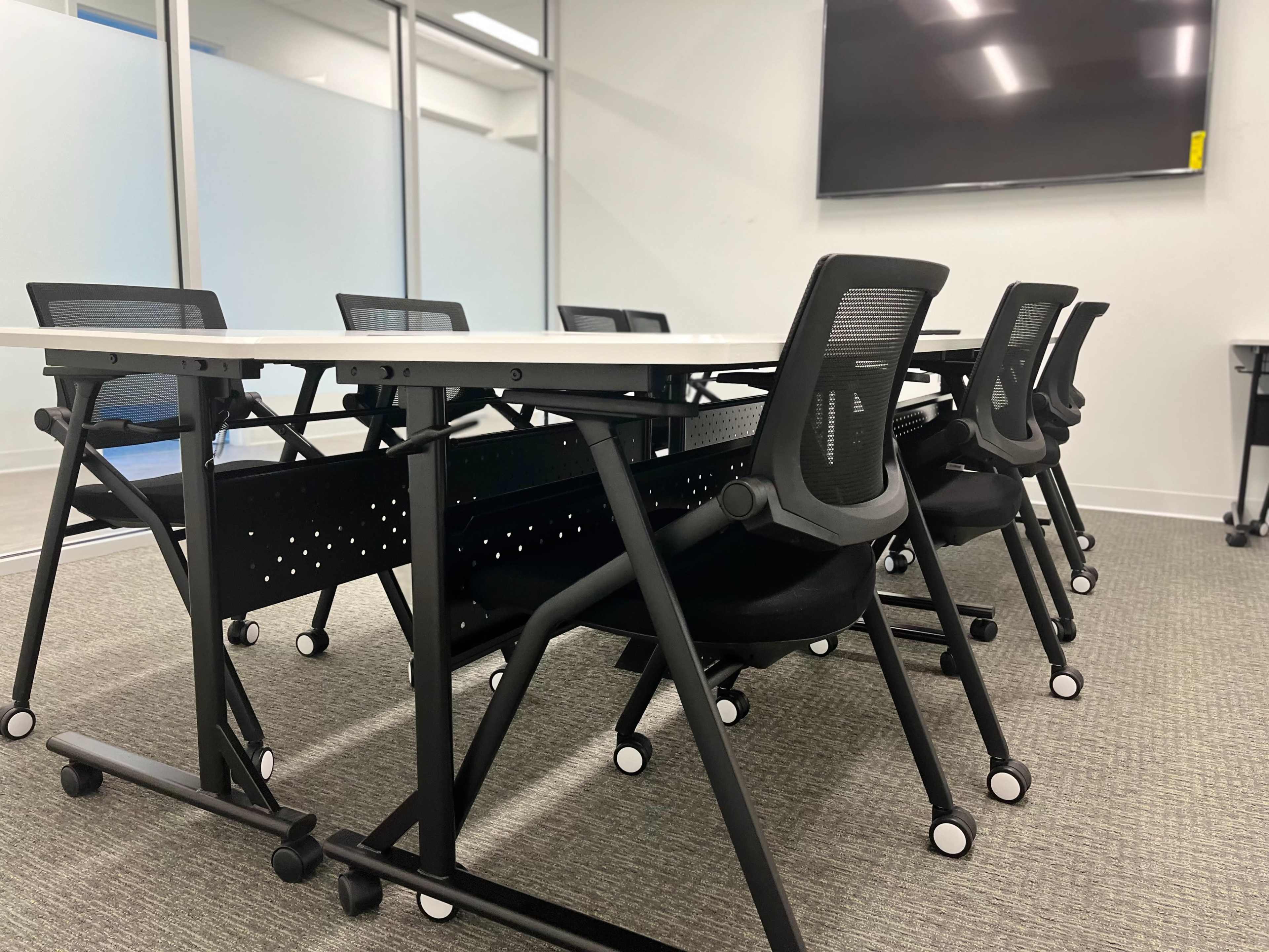 A modern meeting room features a row of folding tables and black rolling chairs facing a large display screen.