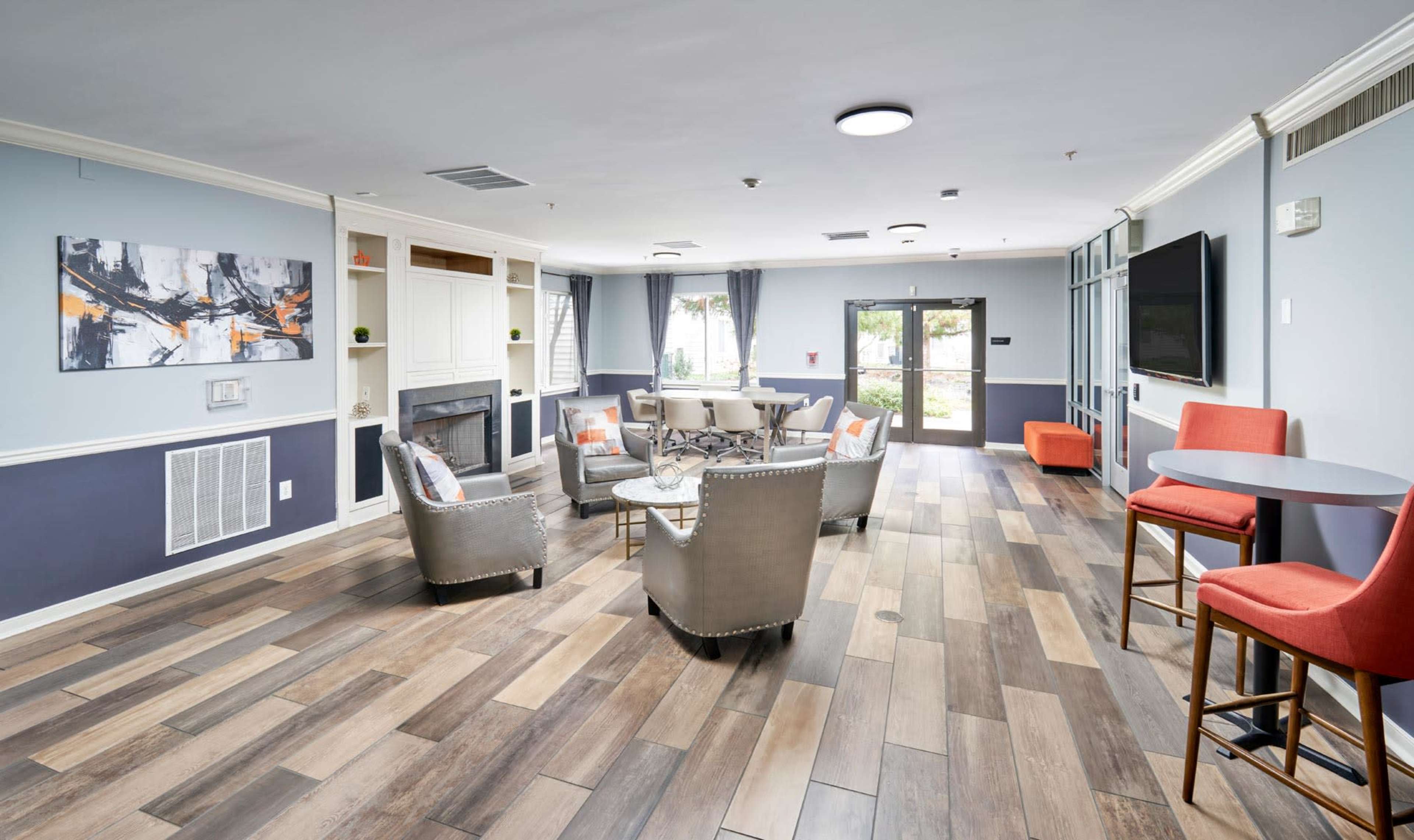 The image shows a well-lit common area featuring a circular table with seating, surrounded by chairs and a couch, along with a bar table and a wall-mounted television.