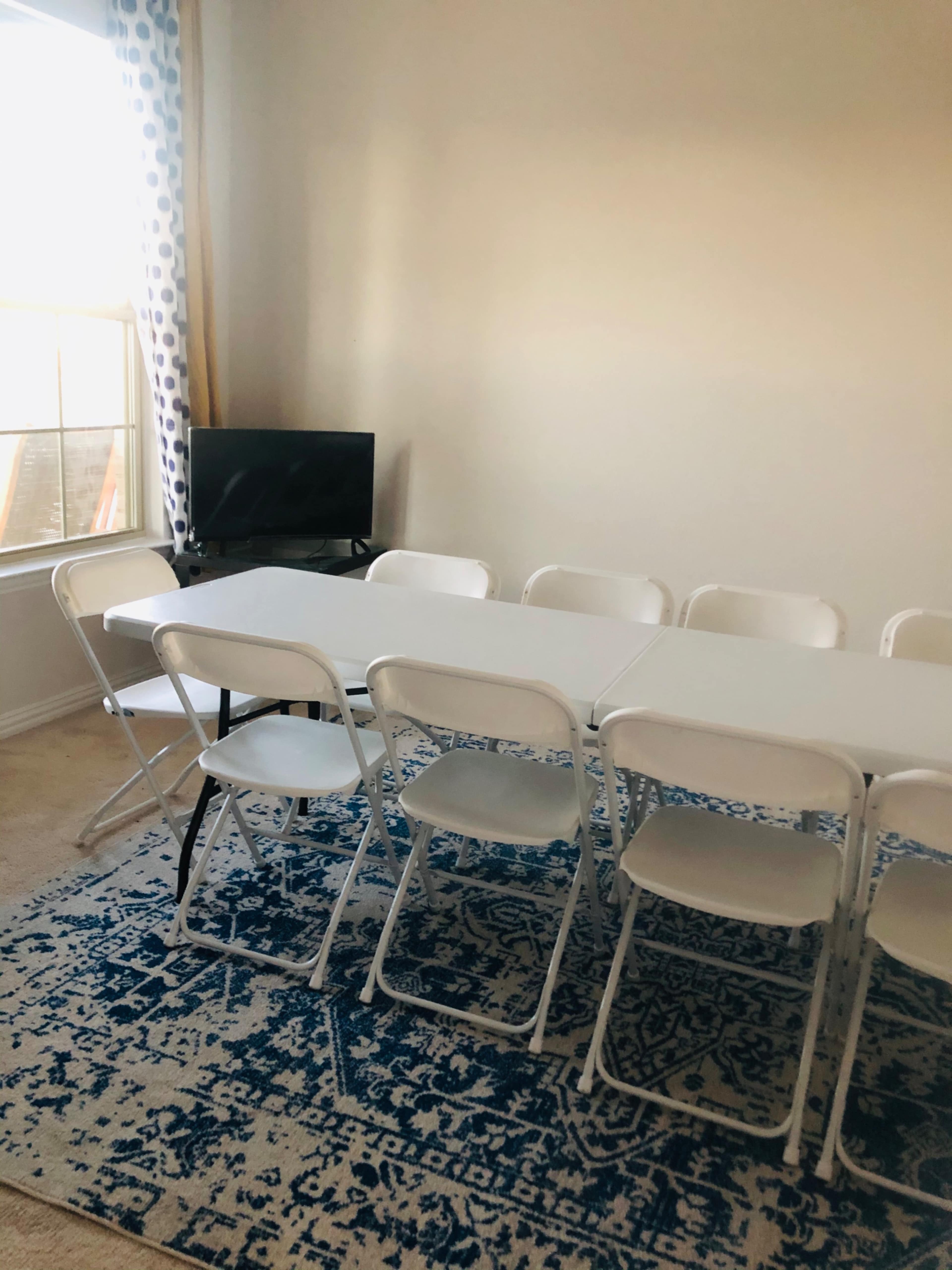 A rectangular table with white folding chairs is arranged on a blue patterned rug in a well-lit room with a window and a small television.