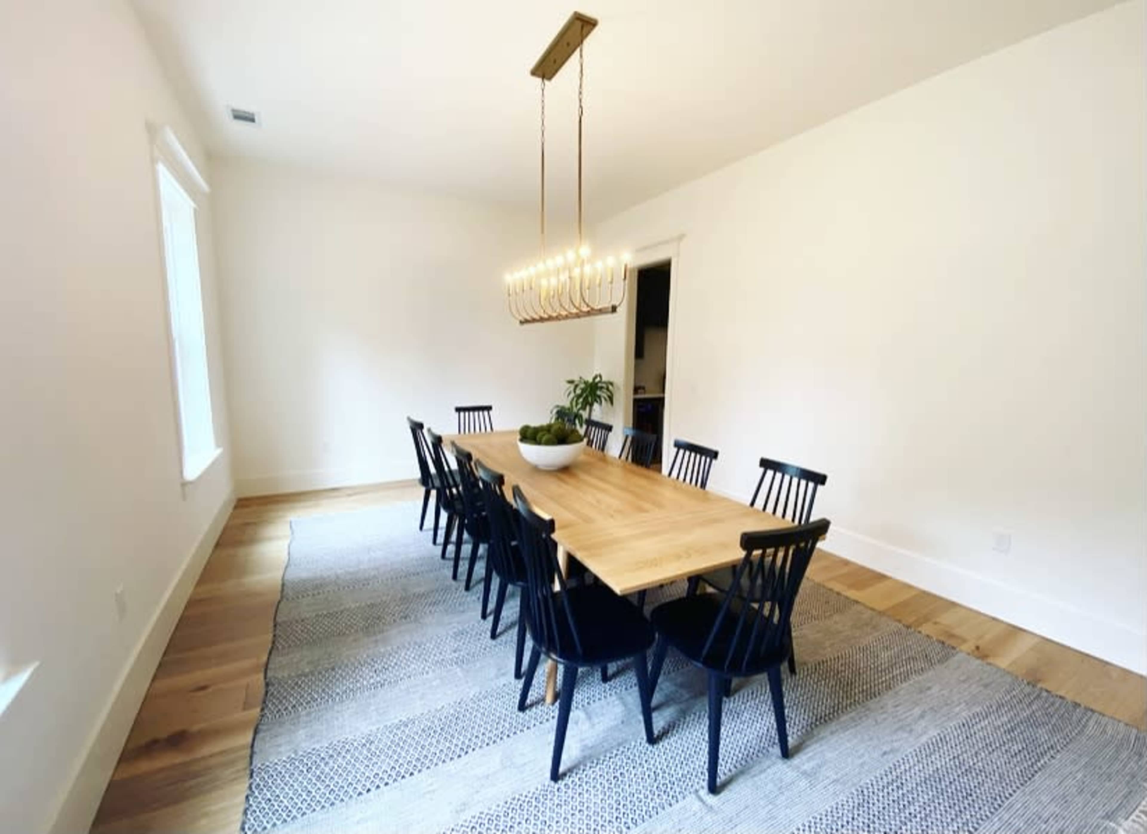 A spacious dining room features a large wooden table surrounded by black chairs, with a chandelier overhead and a plant in a bowl as a centerpiece.