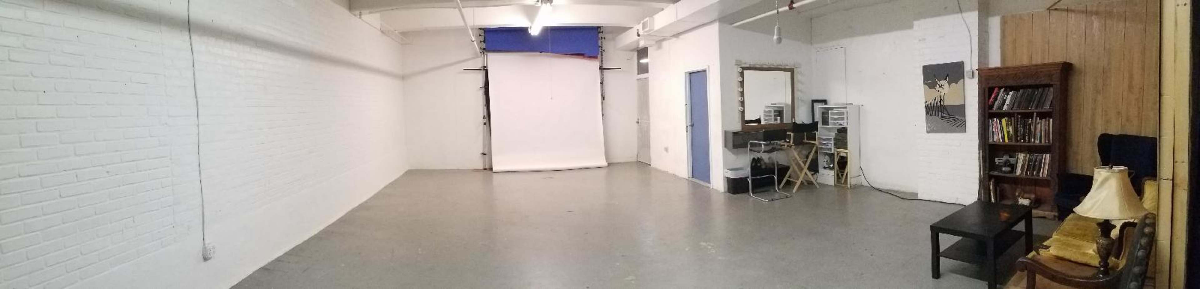 A spacious, empty studio with white brick walls, a gray floor, and minimal furniture including a table, a lamp, and a bookshelf.