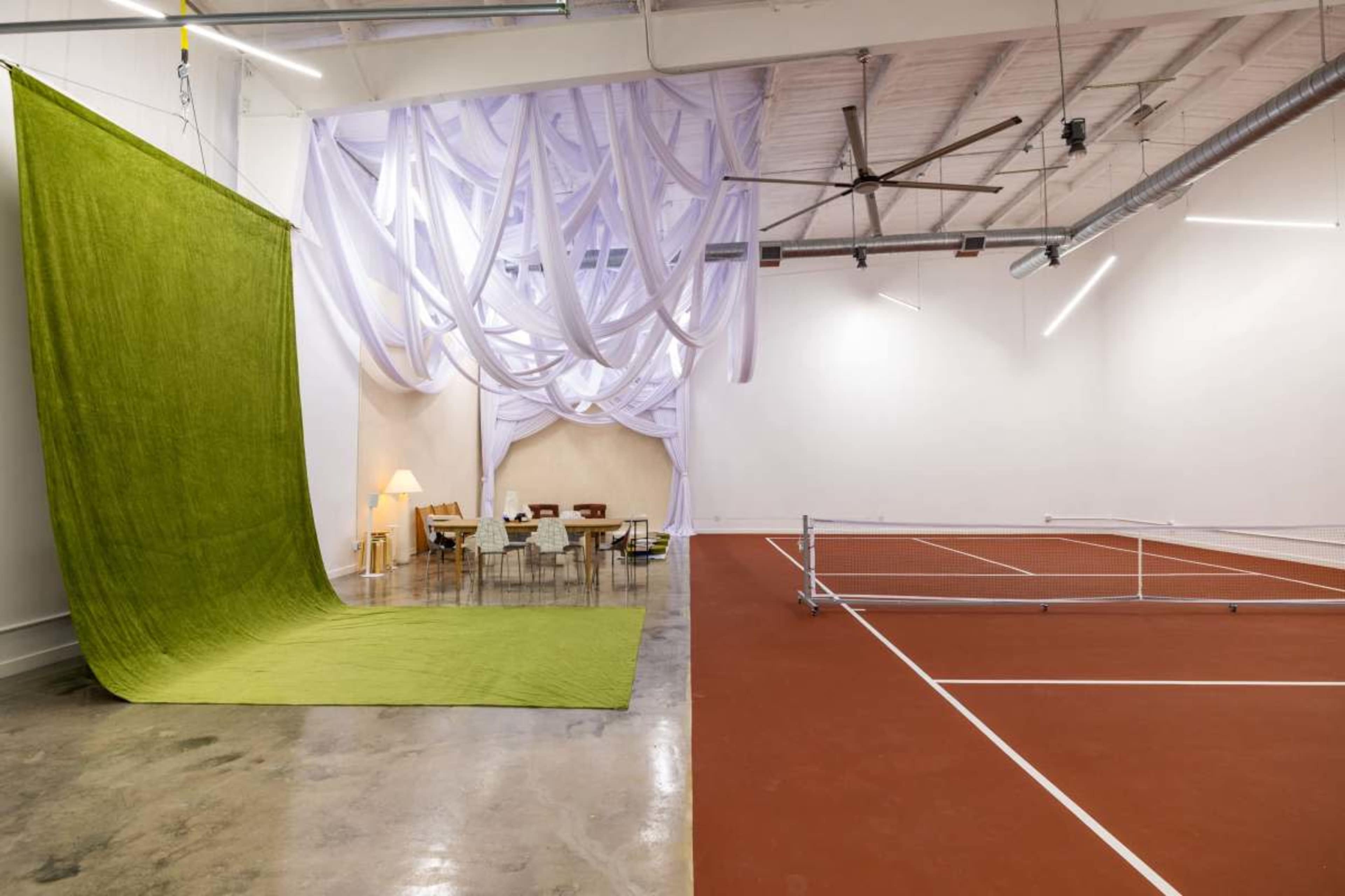 The image shows a spacious room divided into two areas, one featuring a large green fabric backdrop and the other outfitted with a tennis court surrounded by white lines.