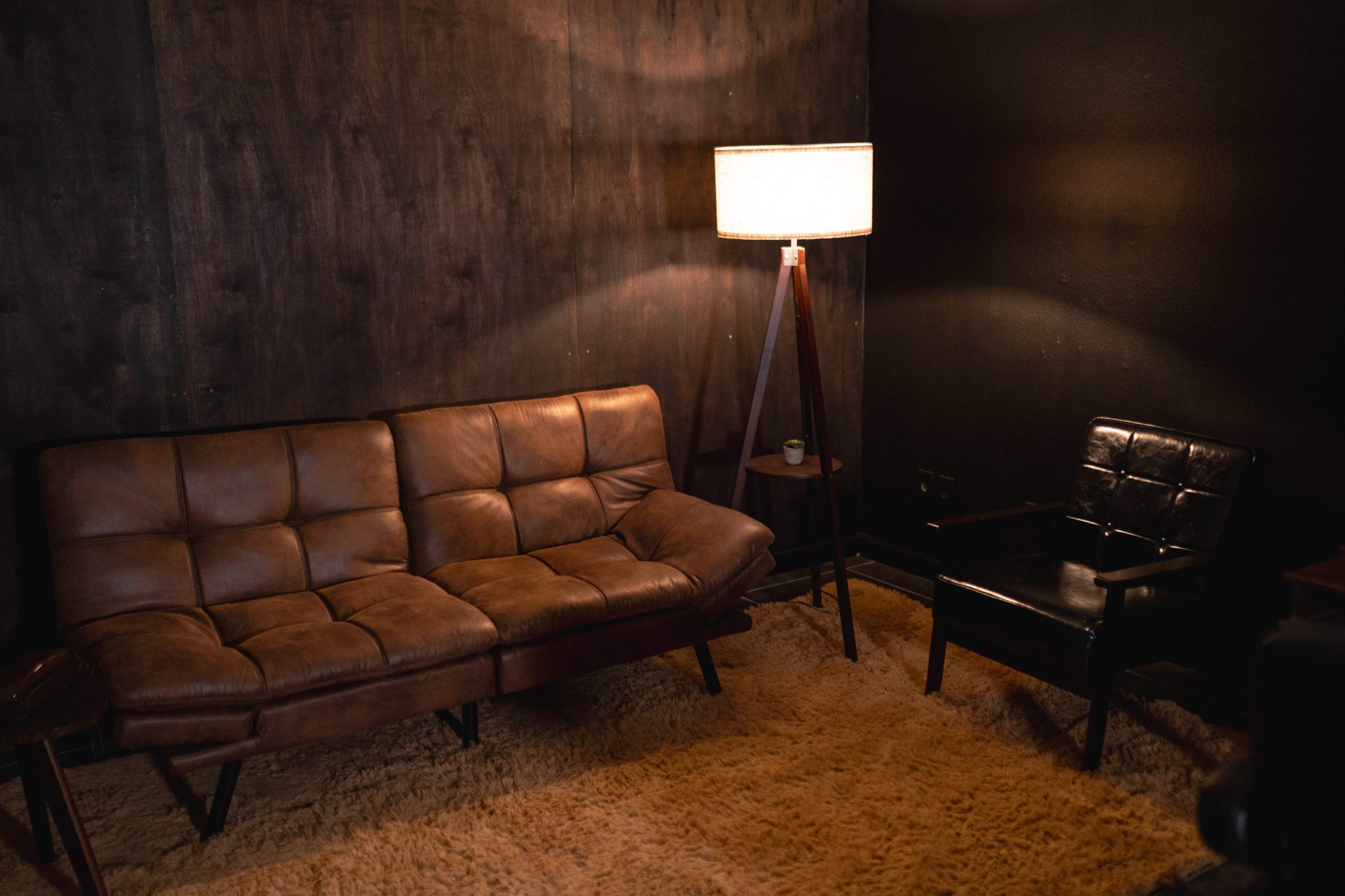 A brown leather sofa and a black armchair positioned in a room with dark wooden walls and a standing lamp providing soft light.