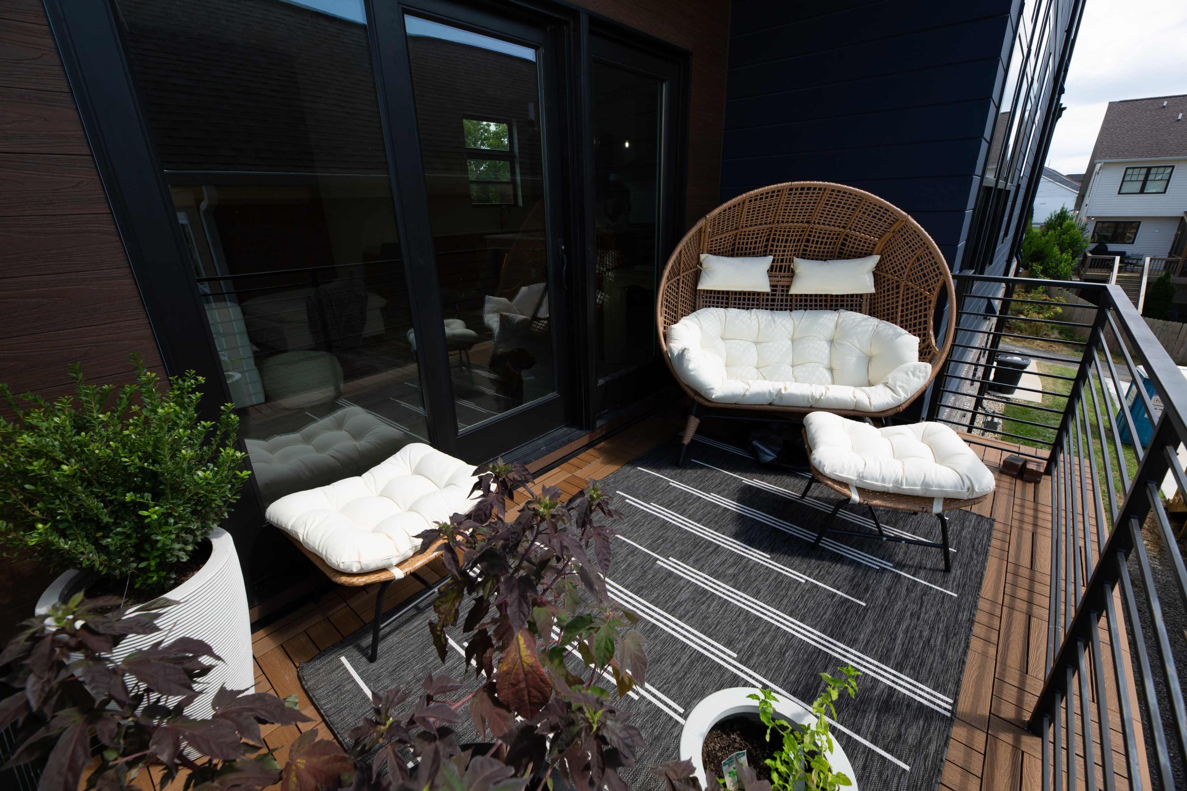 The image shows a balcony with two tufted white chairs and a round wicker chair, surrounded by potted plants and a striped rug.