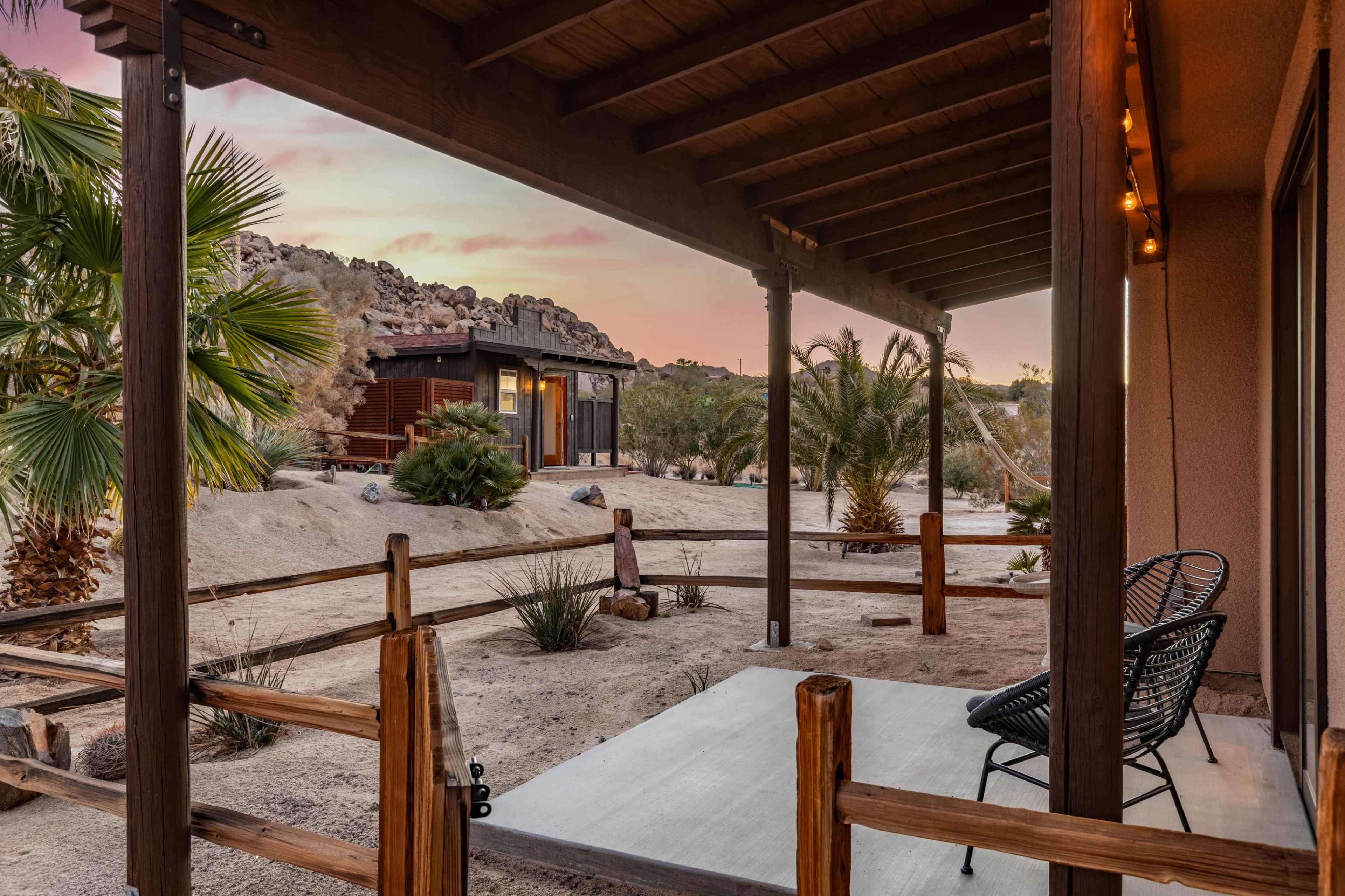 A porch with two chairs overlooks a desert landscape featuring palm trees and rocky outcrops at sunset.