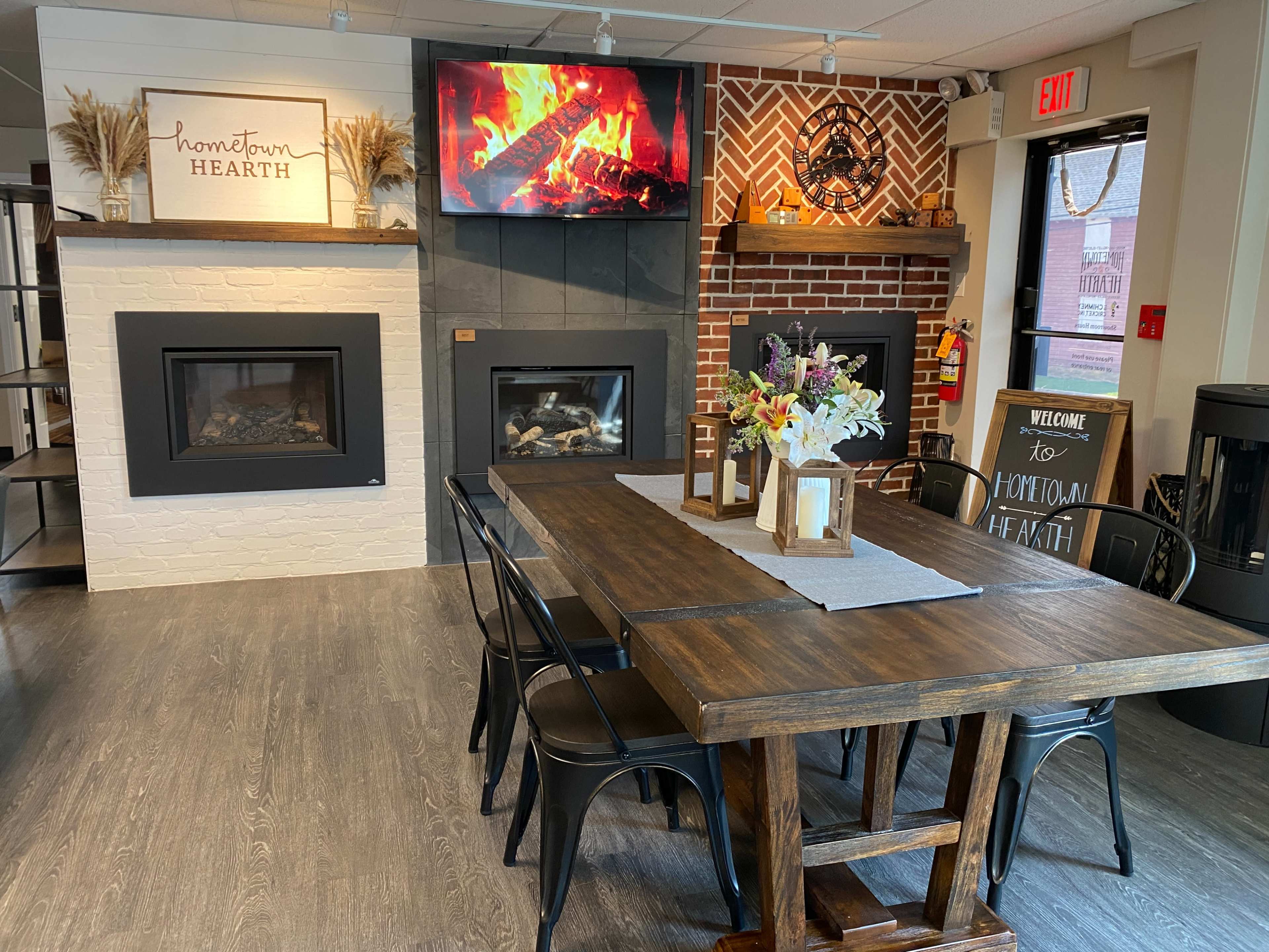 A cozy showroom with a dining table surrounded by metal chairs, fireplaces on display, and a large television showing a fireplace scene.