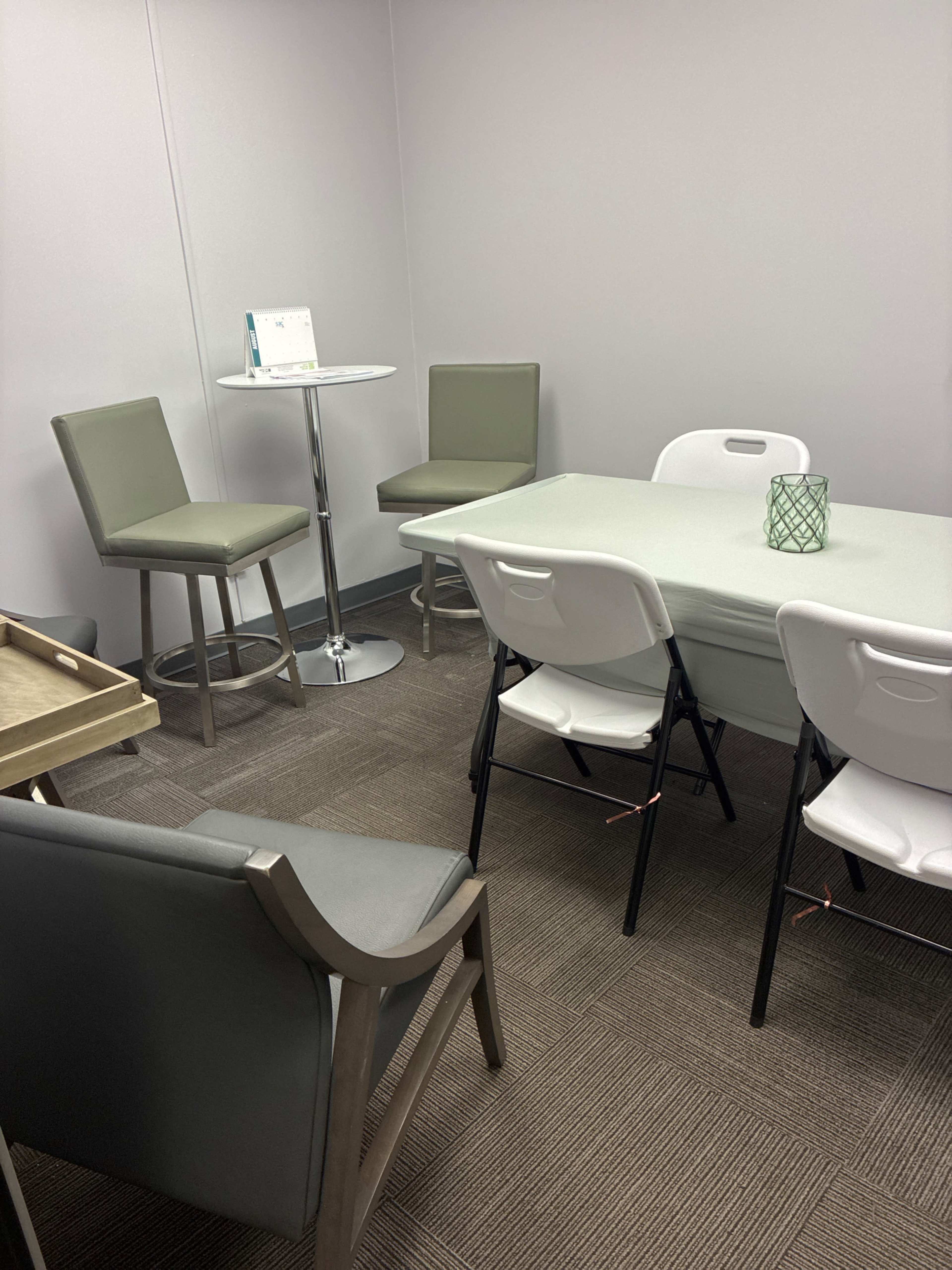 The image shows a small meeting room with a round high table, a rectangular table, and several chairs arranged around them.