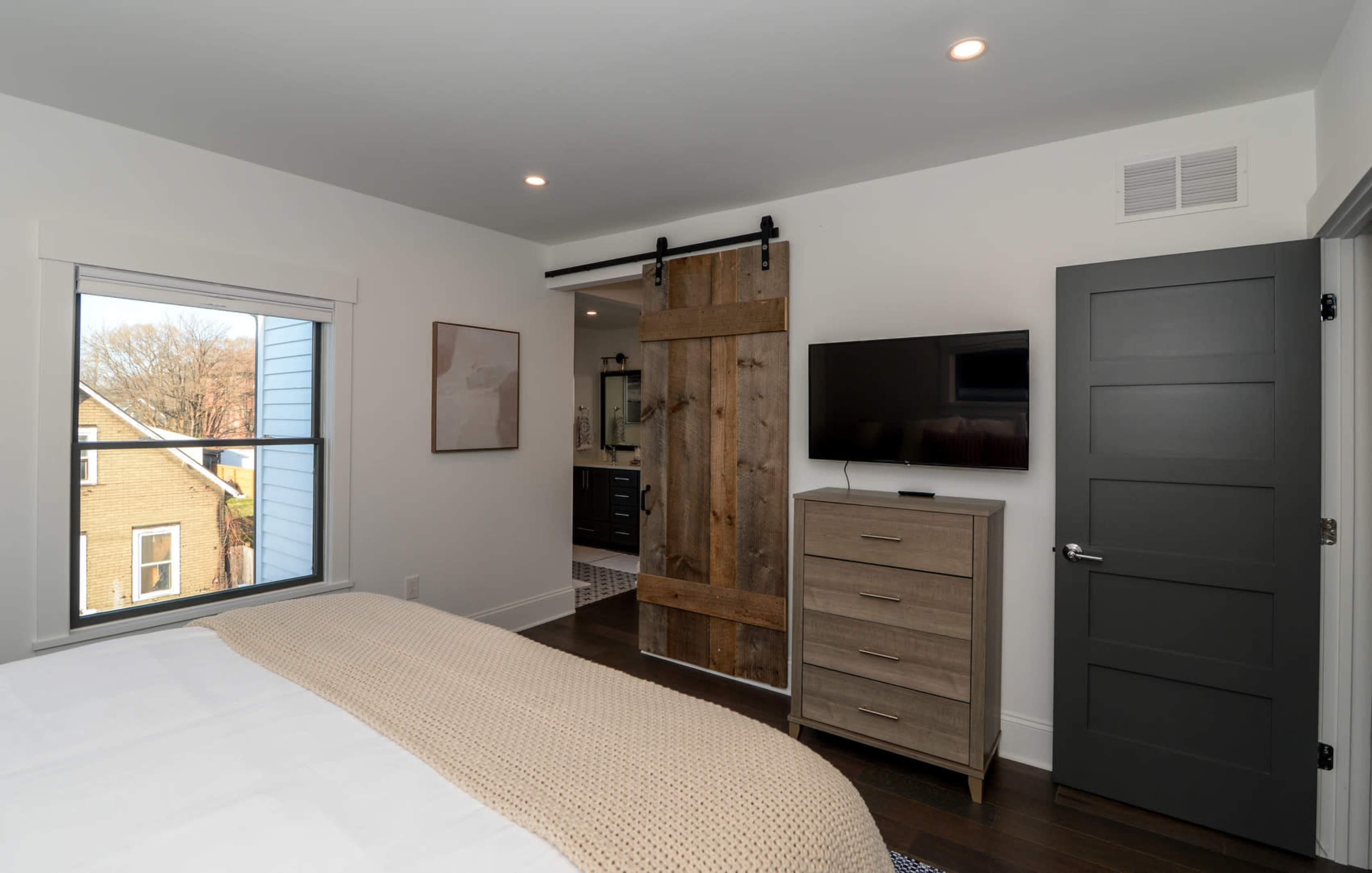 A modern bedroom features a bed with a neutral blanket, a dresser, a flat-screen TV on the wall, and a sliding barn door leading to a bathroom.